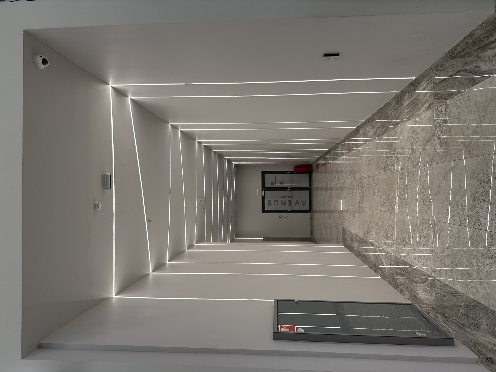 The image shows a well-lit hallway with distinctive linear lighting embedded in the ceiling and walls. The hallway appears to be in a modern building with gray marble and white surfaces. At the end of the hallway, there is a door with the word "AVENUE" on it, written upside down. A security camera is visible in the upper left corner. A window is on the bottom right side of the image. A fire extinguisher is near the end of the hallway. The location could possibly be in Moscow, Russia, but this cannot be confirmed from the image itself. There are no people visible in the image.