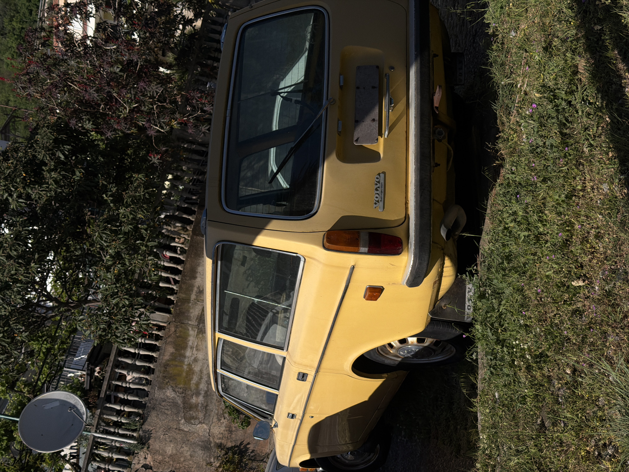 A yellow Volvo station wagon is parked on an overgrown roadside in Stupovi, Montenegro. The vehicle is positioned adjacent to a stone wall and a variety of vegetation, including grass and a tree with dark foliage. A satellite dish is visible in the background, attached to a building. The scene appears to be taken in daylight.