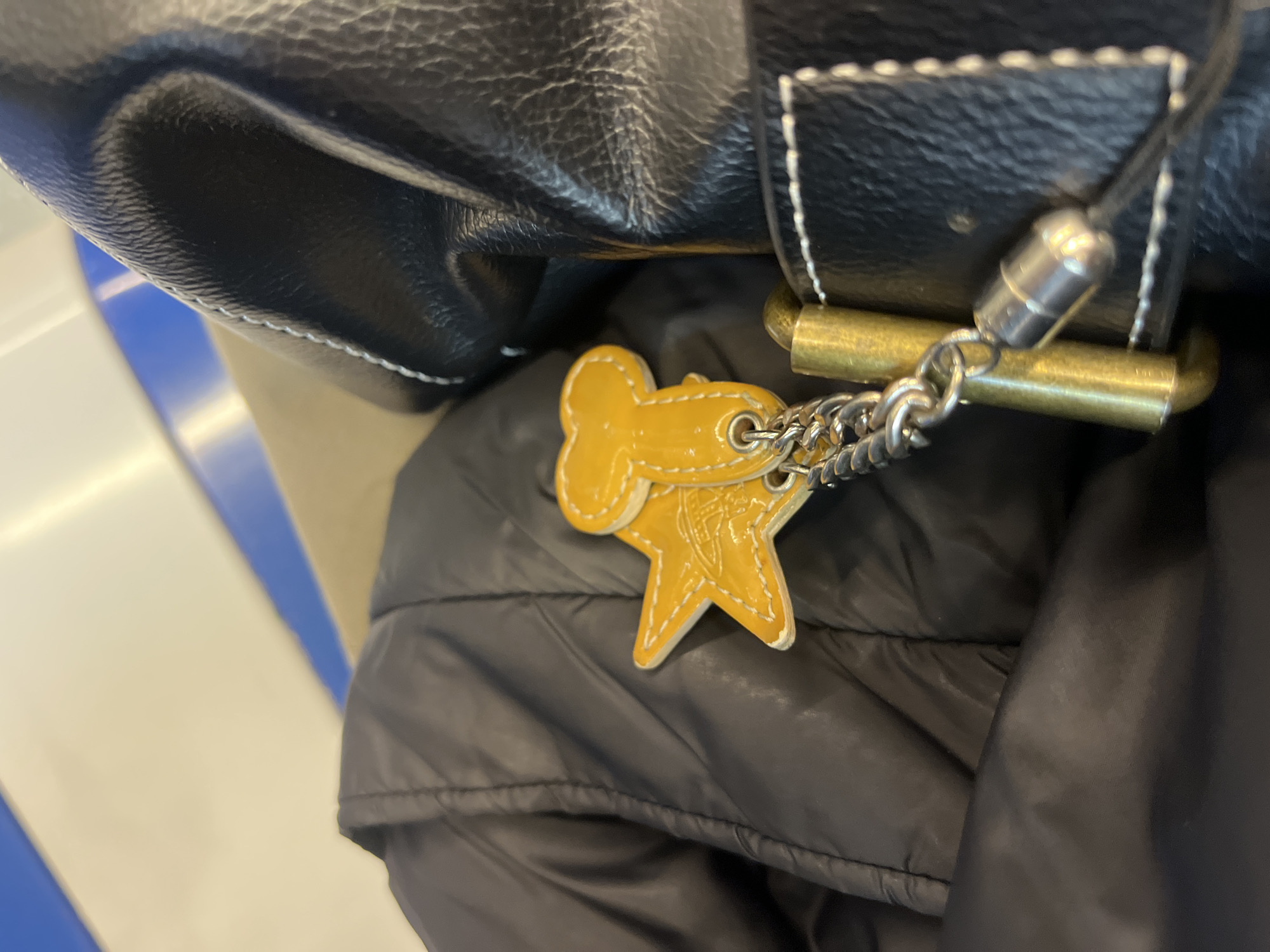 The image presents a close-up view of personal belongings, primarily a black leather bag and a keyring, resting on a black puffy jacket. The black leather bag, visible in the upper portion, features distinctive white stitching along its edges and a slightly textured finish. Attached to the bag's hardware, which includes a brass-colored bar, is a silver-toned chain connected to a keyring.
The keyring holds two bright yellow charms, both appearing to be made of a shiny, possibly enameled or patent leather material with white stitching. One charm is shaped like a dog bone, while the other is a five-pointed star. The star-shaped charm notably displays an embossed design resembling the Vivienne Westwood orb logo, characterized by a ringed planet with a cross at its apex.
Below the bag and charms, a segment of a black puffy jacket is visible, showcasing its quilted texture. The background is out of focus, revealing blurred shapes of a bright white surface and a blue stripe, suggesting an indoor setting such as public transport or a waiting area. The lighting appears artificial. The location context provided is Barcelona, Spain, although the image itself does not contain specific visual cues to confirm this. No other text is discernible in the image.