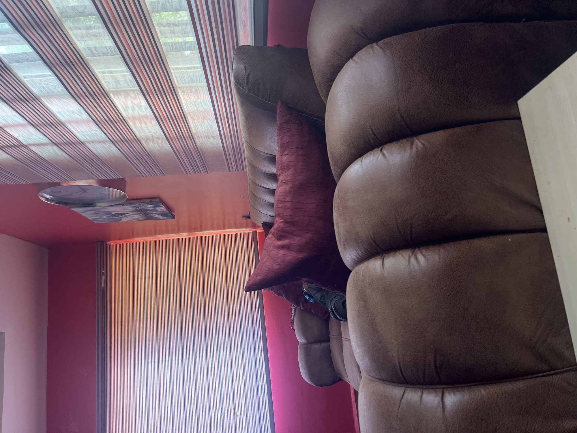 The image shows a close-up, slightly tilted view of a section of a brown leather couch. The couch has plush, cushioned sections, and a deep red decorative pillow with a textured pattern is placed on it. To the left of the couch, a wall painted in a vibrant red is visible, with a striped curtain or blind covering a window or doorway. Above the couch and to the left, a portion of a ceiling with horizontal stripes in shades of brown, white, and red can be seen. A round, metallic object, possibly a clock or a decorative plate, hangs on the red wall. To the far left, a sliver of a lighter pink wall is present. The scene appears to be indoors, in a living space. The lighting suggests it might be daytime, possibly late afternoon due to the warm tones of the light filtering through the blinds. There are no people visible in the image. The overall impression is of a comfortable, albeit slightly dated, interior.