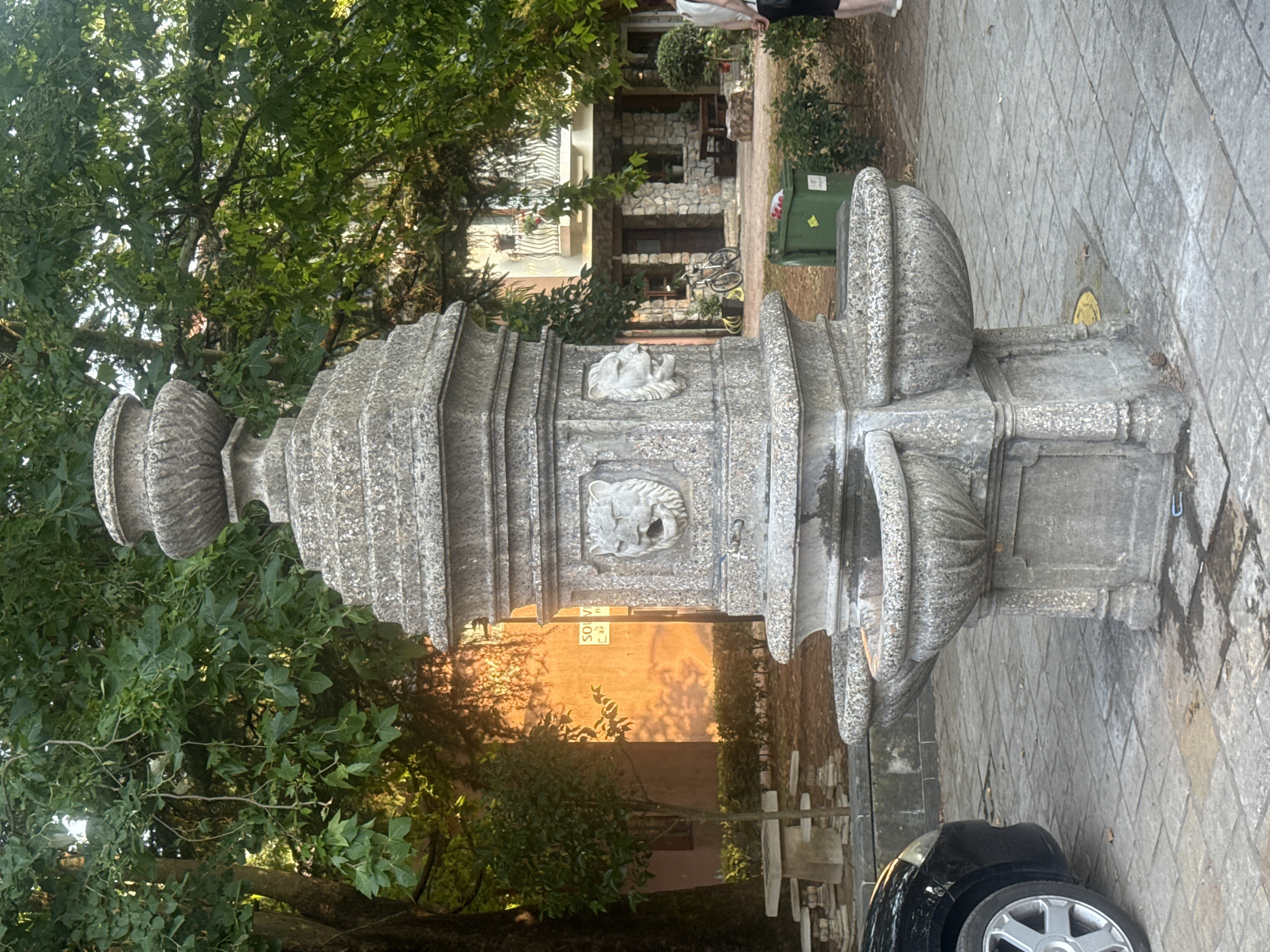 The image shows a decorative stone fountain in Virpazar, Montenegro. The fountain is the central subject, featuring two lion heads carved into its structure, from which water appears to have once flowed. It has multiple tiers and ornate carvings, with two basin-like structures at the lower level. The fountain is situated on a paved area, likely a public square or walkway. In the background, lush green trees and foliage create a natural setting. A building with stone detailing and a balcony is visible behind the fountain, suggesting a residential or commercial area. A bicycle is parked near the entrance of the building. To the left of the fountain, part of a black car is visible. A green trash bin is located behind the fountain to the right. A woman is partially visible on the far right, facing away from the camera. The overall scene depicts a tranquil outdoor setting, possibly a town center or a park area within Virpazar.