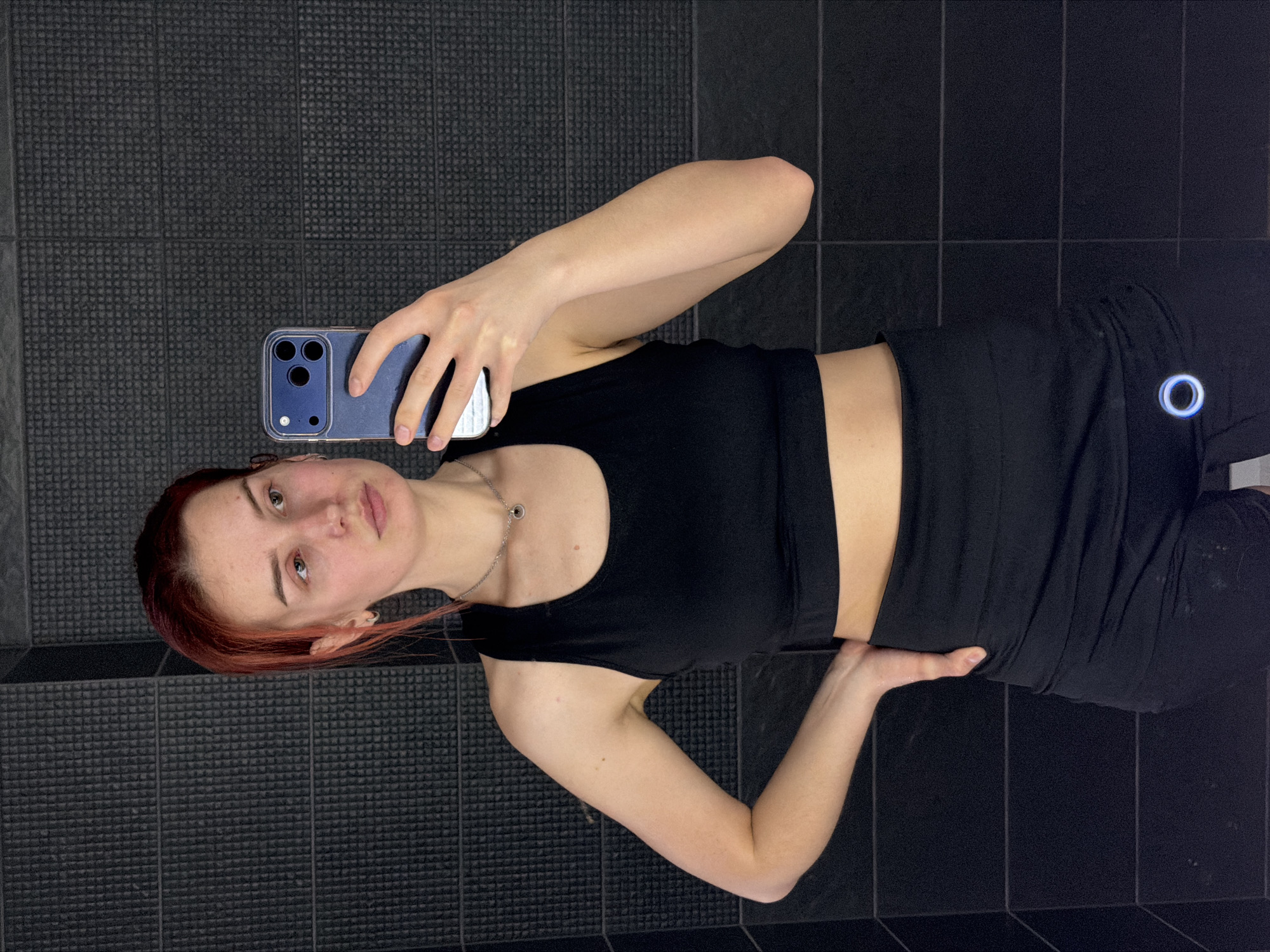 A young woman is taking a selfie in what appears to be a bathroom or gym changing area. She is wearing a black sports bra and black leggings. Her hair is reddish-brown. She is holding a blue iPhone in her left hand, using it to take the picture. The background consists of dark gray and black tiles. A logo is visible on her leggings. The location is not identifiable as the city of Chaikovsky, Russia based on the visual information.