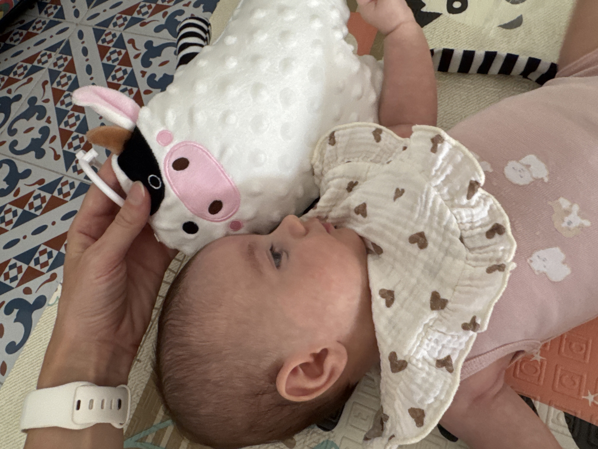 A baby is lying on its side on a mat patterned with colorful geometric shapes. The baby is wearing a pink outfit and a white bib with brown heart patterns. Next to the baby is a plush toy cow. A person's hand is gently touching the cow's head. The scene is an indoor environment, likely a home. There is no information in the image to suggest the location is Isola delle Femmine, Italy.