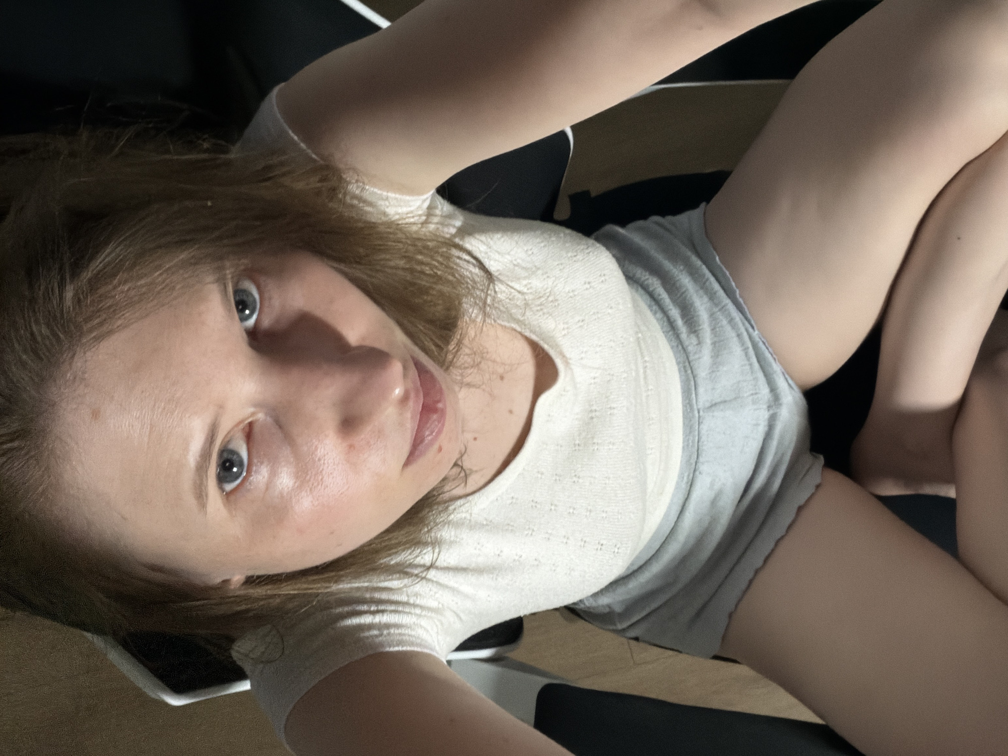 A young Caucasian woman with blonde hair and blue eyes is seated in a chair, looking directly at the camera. She is wearing a white short-sleeved top and gray shorts. The background is dark and appears to be indoors, with some lighting creating shadows on her face and body. The image is a close-up shot, focusing on the upper half of her body.