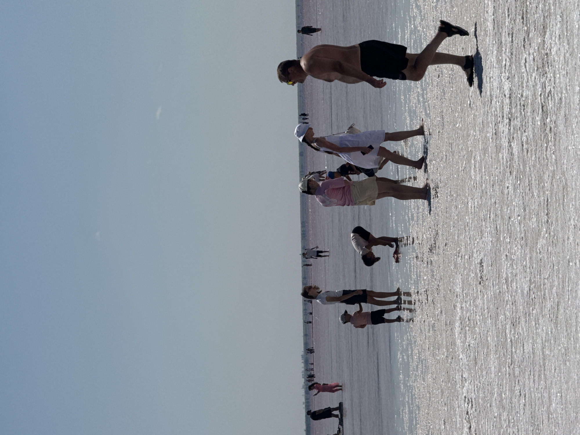 The image shows a wide, shallow body of pink water, likely a salt lake. The ground is covered in a crystalline salt crust. In the foreground, several people are walking or standing in the water. A man in black shorts and sandals is walking towards the right. To his left, a woman in a white dress and hat walks away, and a man in a white shirt is partially visible. Further back, a woman in a pink t-shirt and shorts bends down, and a child in a blue shirt and dark shorts stands nearby. On the left side of the frame, a woman in a pink dress stands, and a man in dark shorts is visible near the edge. In the distance, more people are scattered across the lake. The sky is clear and blue. The location is identified as the city of Bursol', Russia.
