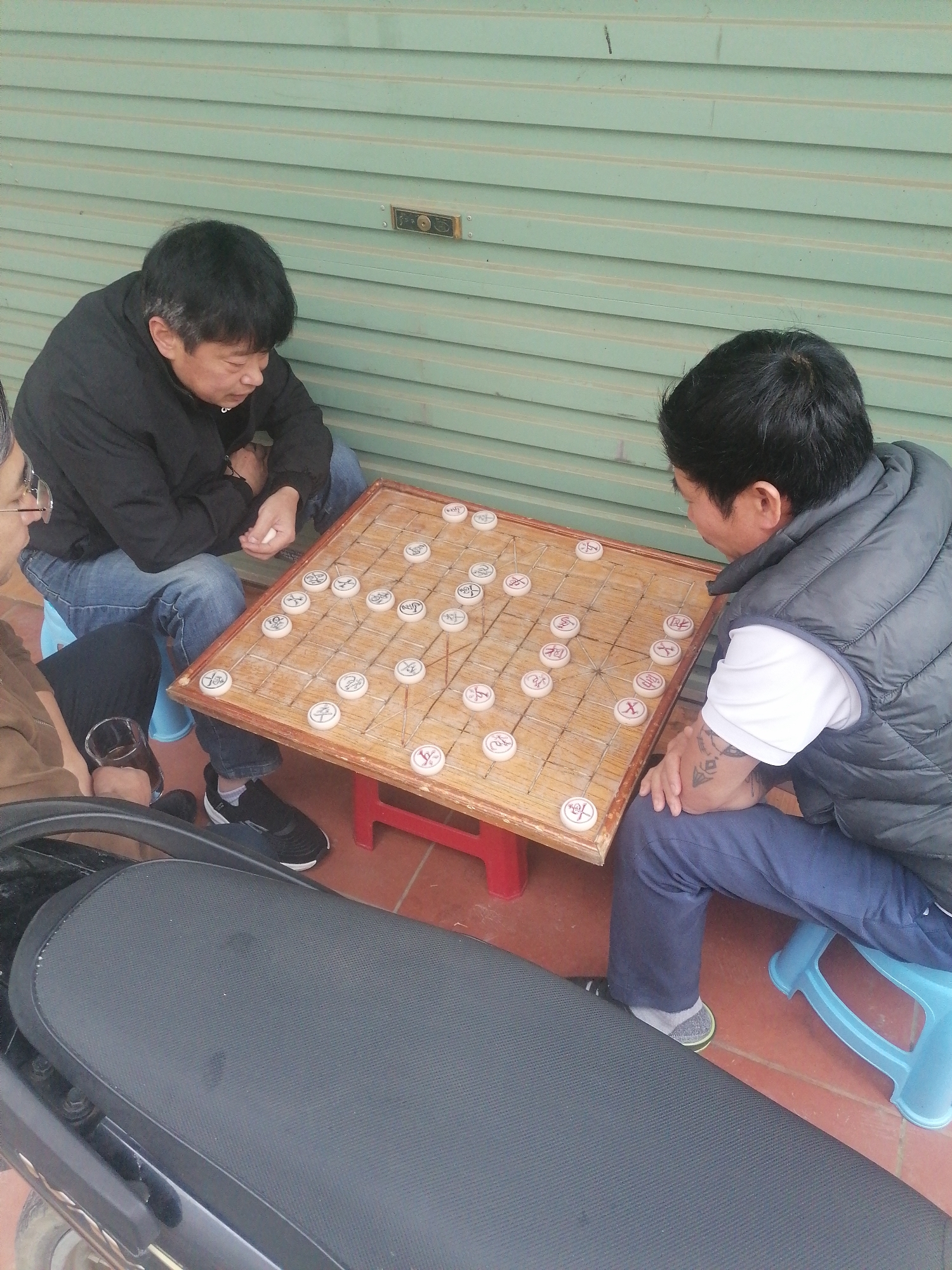 Three men are gathered around a wooden board game, engaged in a casual outdoor activity, likely in Sa Pa Ward, Vietnam, as indicated by the location context. Two of the men are actively playing what appears to be Chinese Chess (Xiangqi), with the board resting on a small red stool between them. Both players are seated on small blue plastic stools. The man on the left is wearing a black jacket and blue jeans, intensely focused on the game. His hands are clasped, and he appears to be contemplating his next move. The man on the right, wearing a dark vest over a white t-shirt and blue trousers, also looks absorbed in the game. He has a tattoo visible on his left forearm.
A third man is partially visible on the far left, observing the game. He wears a brown jacket and eyeglasses and holds a glass containing a dark beverage, possibly tea. In the foreground, part of a black motorcycle seat is visible, suggesting they might be parked or resting near a vehicle.
The setting is an outdoor or semi-outdoor space, possibly a covered porch or sidewalk. A light green corrugated metal wall, resembling a shop shutter or garage door, forms the background. The ground is covered with reddish-brown tiles. The game pieces are round and white, with distinct Chinese characters visible on them, such as "帥" (general), "馬" (horse), "砲" (cannon), "兵" (pawn), and "卒" (soldier). The overall atmosphere is relaxed and contemplative, indicative of a leisurely pastime. The natural lighting suggests it is daytime.