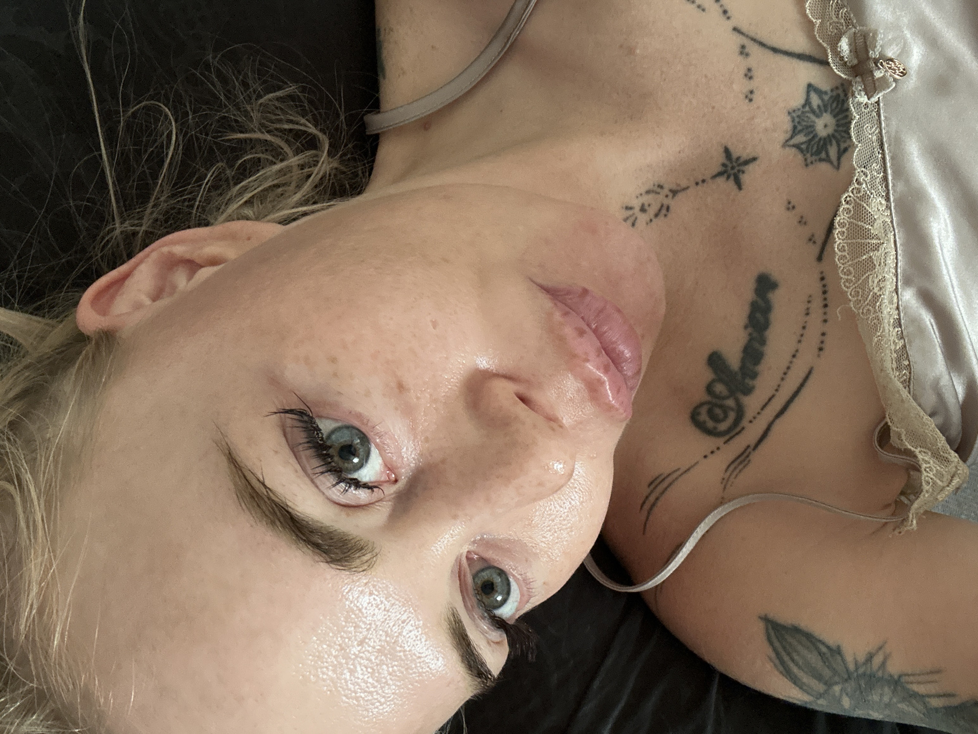 A close-up shot shows a woman with light skin and blonde hair. She is lying down with her head slightly tilted, looking up. Her eyes are open and her gaze is directed towards the viewer. She has fair skin with light freckles across her cheeks and nose. Her eyebrows are well-defined, and she is wearing mascara and possibly false eyelashes. Her lips are slightly parted, and she is wearing a light-colored, possibly satin, camisole. There are tattoos visible on her chest and shoulder. The background is dark and out of focus, suggesting an indoor setting, possibly a bedroom. There are no distinct landmarks or environmental details that would indicate a specific location within a city.