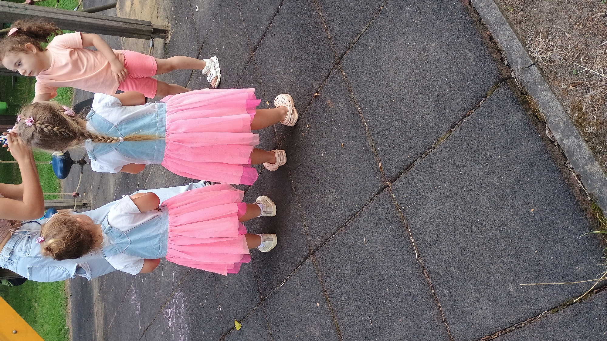 In Sokolov, Czechia, two young girls in matching pink tulle dresses over denim tops and white t-shirts stand on a rubber playground surface. The girl on the left has her hair in a bun with a pink accessory, while the girl on the right has a braid with a pink accessory. They are facing away from the camera, towards a playground structure with a blue element. Another young girl in a peach t-shirt and pink shorts stands to the right, looking at the braided girl. A woman's hands are visible in the background, near the braided girl's head, suggesting she is interacting with her hair. The ground is made of dark gray interlocking rubber tiles, with some grass and dirt visible at the edges.