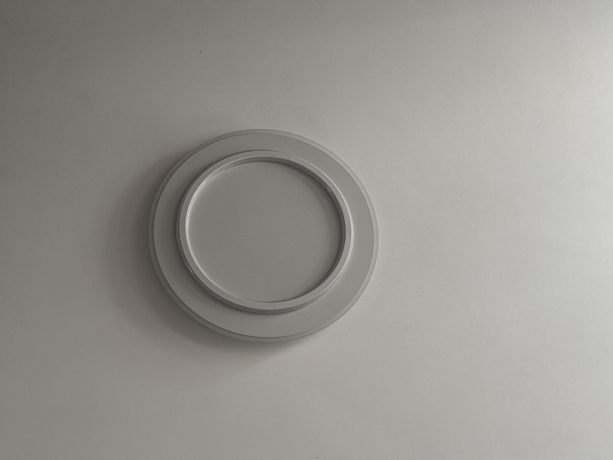 The image shows a close-up, overhead view of a recessed ceiling light fixture. The fixture is circular and has a layered design with a matte white outer ring and a slightly recessed, lighter gray inner circle. The surrounding ceiling is a uniform, light gray color, with subtle shadows cast by the fixture, indicating the light source is overhead. There is no indication of the location being in Vsevolodovo, Russia, or any other specific city. The scene is static and devoid of any people or actions.