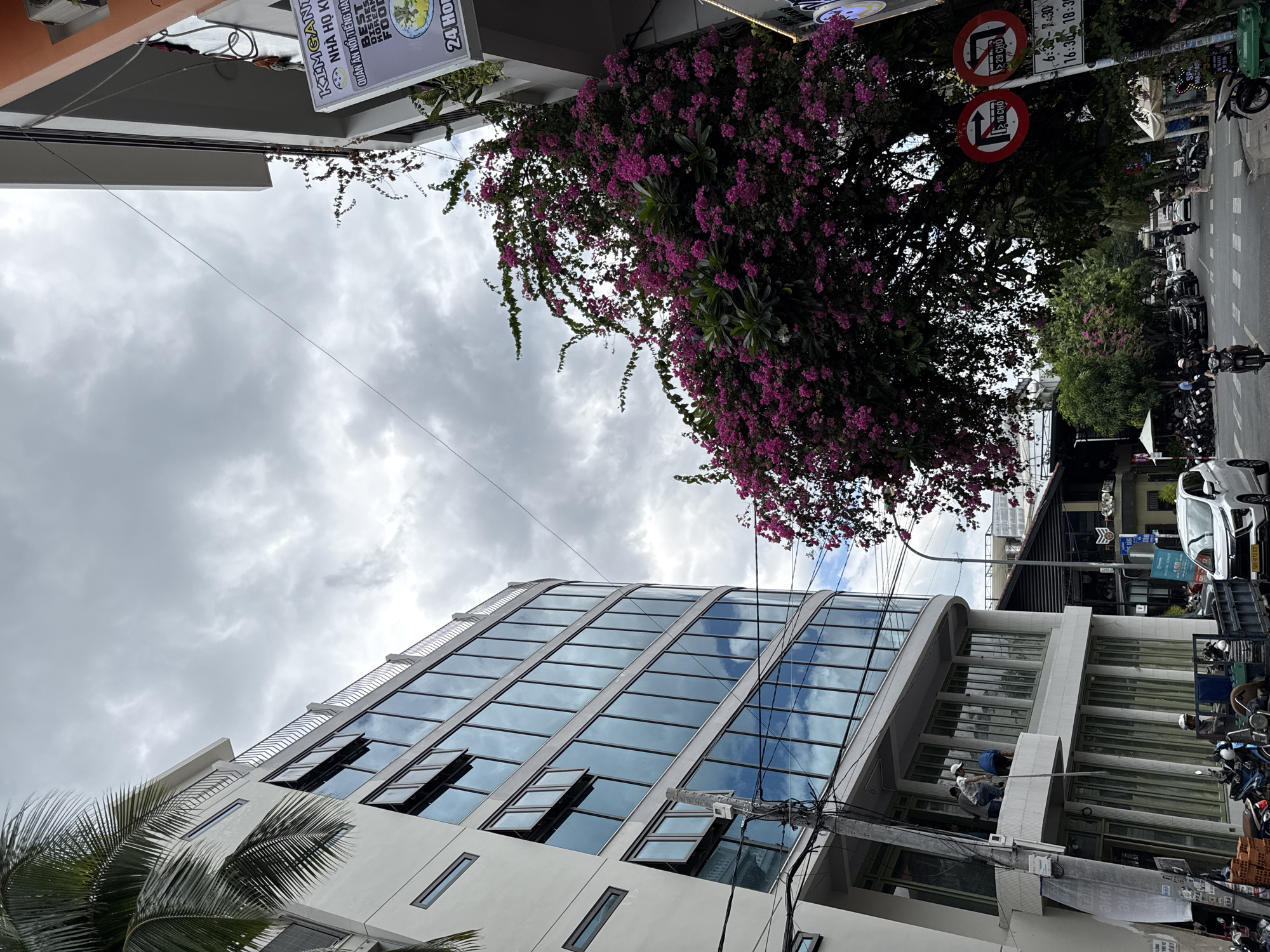 This is a photograph taken from street level, looking upwards at a curved, modern building with many windows. The building appears to be white with dark-framed windows that reflect the cloudy sky. In front of this building, a large tree is covered in vibrant pink flowers, with some of its branches extending over the street. To the right of the pink-flowered tree, another building, partially visible, has an orange facade and a sign that reads "KIM GANTU NHA HO KIEN DINH". Below this sign, another sign is partially visible with the text "24 HOURS". The sky is overcast with dramatic, dark clouds.
The street in the foreground is paved and has several motorbikes parked on the sides. A white car is visible driving down the street, and several people are walking or riding motorbikes. There are also traffic signs visible. One sign shows a red circle with a diagonal line and a symbol of a vehicle turning left, with text below indicating traffic restrictions. Another sign has similar red circle markings with symbols of vehicles. A timetable sign with times "6:30 - 7:30" and "16:30 - 18:30" is also visible.
On the balcony of the white building, three men are visible, seemingly working or observing. One man is wearing a white helmet and blue shirt, another is wearing a blue shirt and white cap, and the third is wearing a dark shirt.
The scene suggests a commercial street in a city, likely Nha Trang, Vietnam, given the signage in Vietnamese and the prevalent mode of transportation. The architecture and the presence of flowering trees are characteristic of urban environments in Southeast Asia.