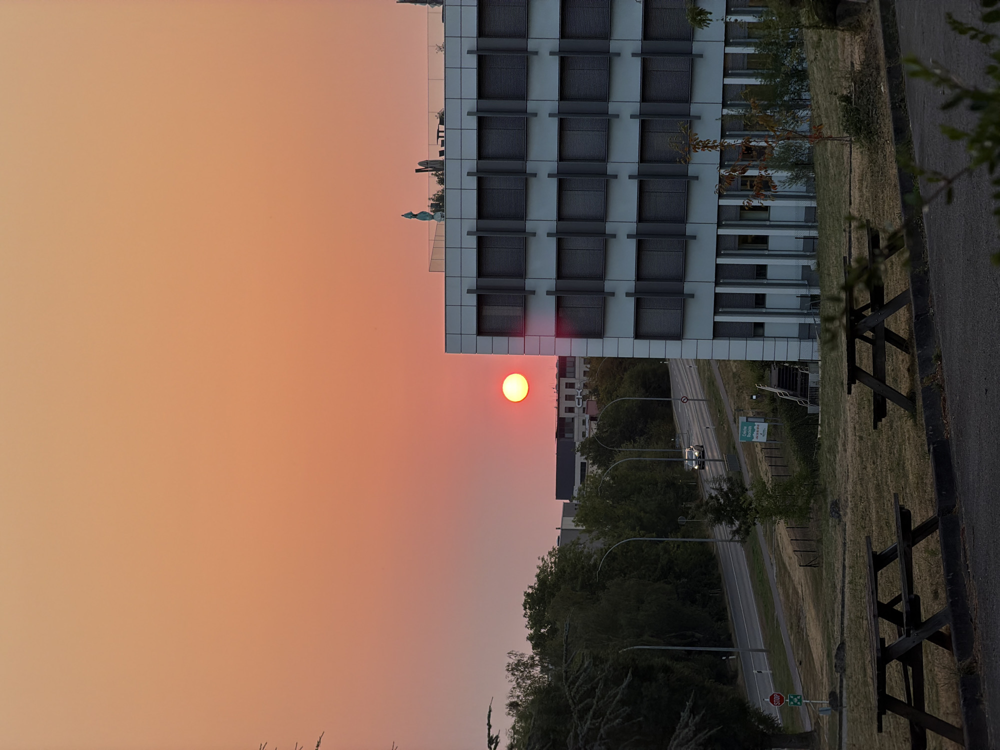 The image depicts a sunset over an office building in Leudelange, Luxembourg. The sky is a gradient of orange and pink, with the sun a bright red orb on the left side. A modern, multi-story office building with rows of closed windows dominates the right side of the frame. In the foreground, two wooden picnic tables are visible on a patch of dry grass. A road with a few cars driving on it is visible behind the building, lined with trees and streetlights. A stop sign is also present near the road. The scene appears to be set in a business or industrial area on the outskirts of the city.