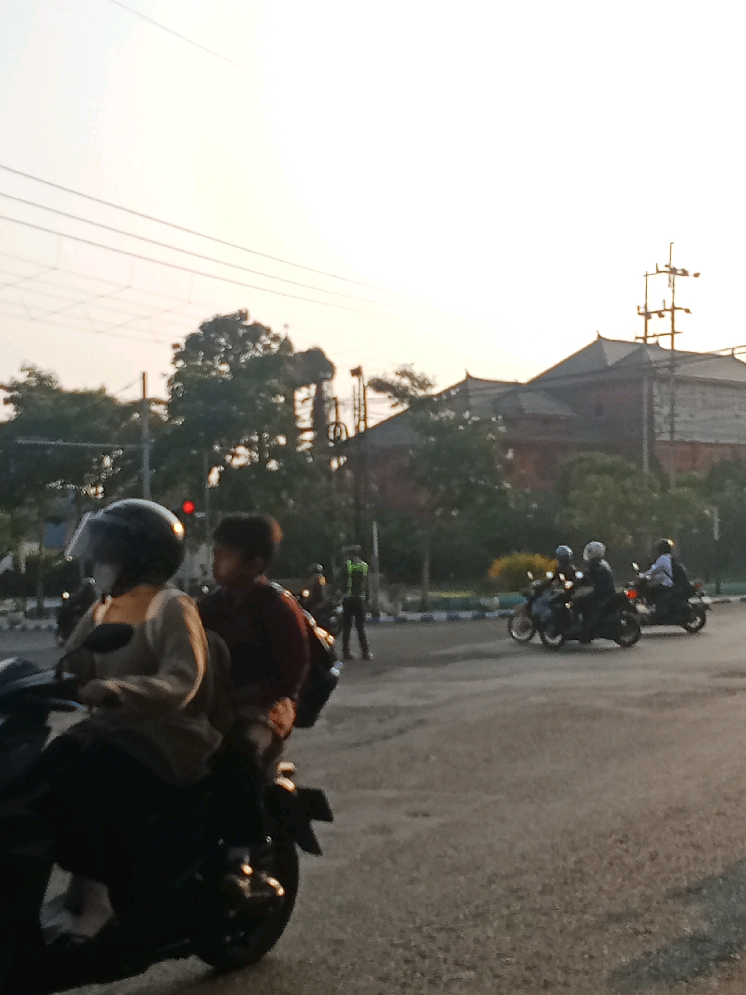 The image captures a street scene in Kaliwungu, Indonesia, likely during the late afternoon or early evening, indicated by the soft, warm lighting. In the foreground, two individuals are riding a motorcycle, with the person in front wearing a black helmet and a light-colored long-sleeved shirt. The person behind them is wearing a dark maroon shirt. They are positioned on a paved road, with other motorcycles and riders visible in the mid-ground and background.
In the mid-ground, a traffic light with a red illuminated signal is visible behind trees. Several motorcycles with riders wearing helmets are proceeding on the road. To the right, two more motorcycles are in motion. In the background, a large, dark building with a distinctive tiered roof, characteristic of Indonesian architecture, stands out against the sky. A tall, utility pole with multiple crossbars is situated near the building. The environment appears to be an urban or semi-urban setting, with trees lining the road and the presence of electrical infrastructure. The overall mood of the image is that of a typical day in a busy Indonesian town.