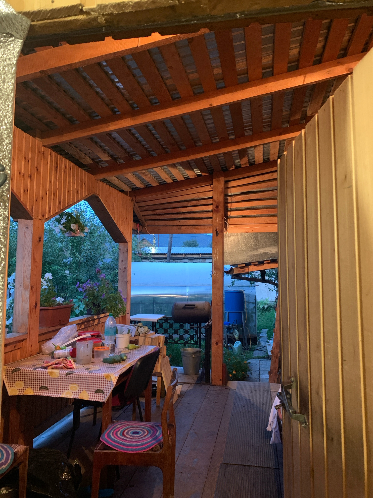 The image depicts a wooden porch in Asha, Russia, likely during the evening. A table covered with a checkered tablecloth is set with various items, including a bottle of water, cups, and food containers. Several chairs surround the table, one of which has a circular crocheted cushion. Beyond the table, a view of a garden with lush greenery and flowering plants is visible. In the background, a greenhouse, a barbecue grill, and blue water barrels can be seen. The porch is covered by a wooden roof with exposed beams. The overall scene suggests a rustic, outdoor setting, possibly a backyard or a dacha.