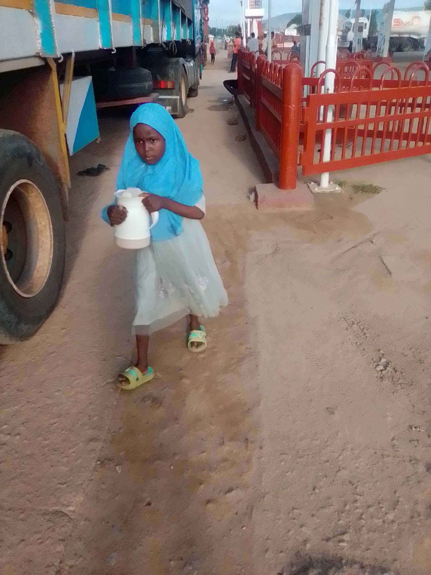 The image depicts a young girl wearing a blue hijab and a light-colored dress, holding a white thermos flask in the city of Kwadon, Nigeria. The girl is standing on a dirt or sand-covered ground, with the side of a large truck visible to her left. In the background, there appears to be a fuel station with red fencing, and other trucks and people are present. The lighting suggests it is daytime.