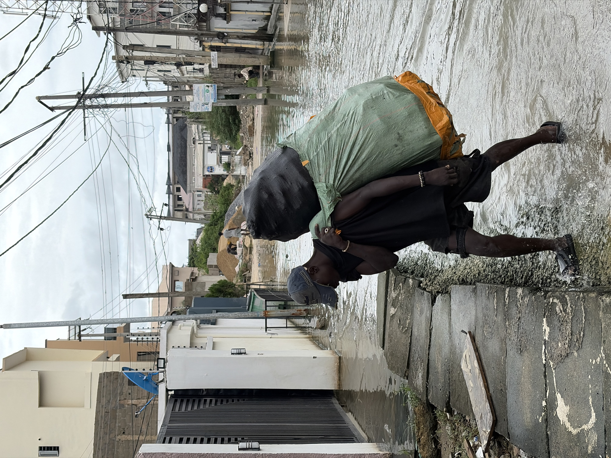 A person is carrying a large sack over their shoulders through flooded streets in Igboefon, Nigeria. The sack is a dark color and is covered by a large, bright green tarp, with an orange flap visible at the top. The person is wearing a dark t-shirt, dark shorts, a cap, and sandals. They are walking on a paved path next to the flooded street. The water is murky and reflects the cloudy sky. In the background, there are several buildings, some of which appear to be multi-story residential homes. There are also numerous utility poles with a complex network of electrical wires crisscrossing overhead. The sky is overcast, suggesting it is daytime, possibly after rain. The scene depicts the challenges of navigating flooded urban environments, likely due to heavy rainfall or poor drainage. The person's posture and the load they are carrying suggest a strenuous effort. Visible text includes signs on poles in the background, but they are not legible.