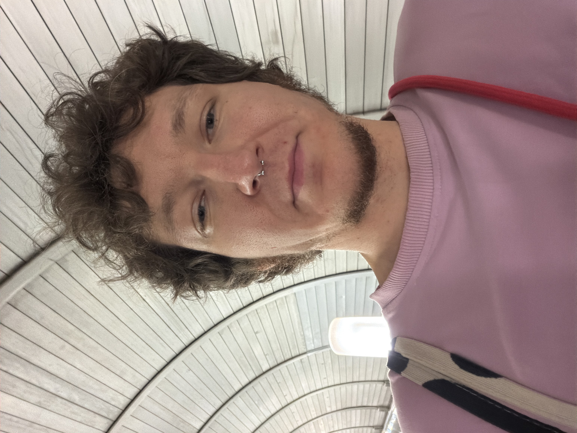 A selfie taken from a low angle shows a person with curly brown hair and a septum piercing. They are wearing a pink crewneck sweatshirt and have a bag strap with a black and white cow print design slung over their left shoulder. A red strap is visible on their right shoulder. The background appears to be an indoor walkway with a curved, ribbed ceiling and a light fixture to the left.