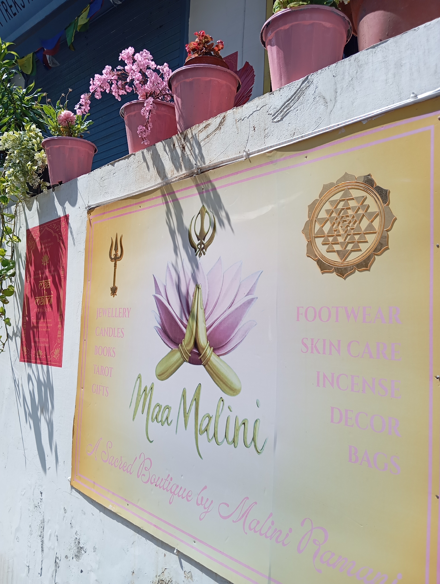 The image shows a sign for "Maa Malini, A Sacred Boutique by Malini Ramani", located on a white-painted wall, presumably in Rishikesh, India, as indicated by a small adjacent sign on the left. The boutique sign lists items for sale like jewelry, candles, books, tarot cards, gifts, footwear, skincare, incense, decor, and bags. The sign includes symbols of a trident, a Khanda (Sikh symbol), and the Sri Yantra. Above the sign, there are pink potted plants.