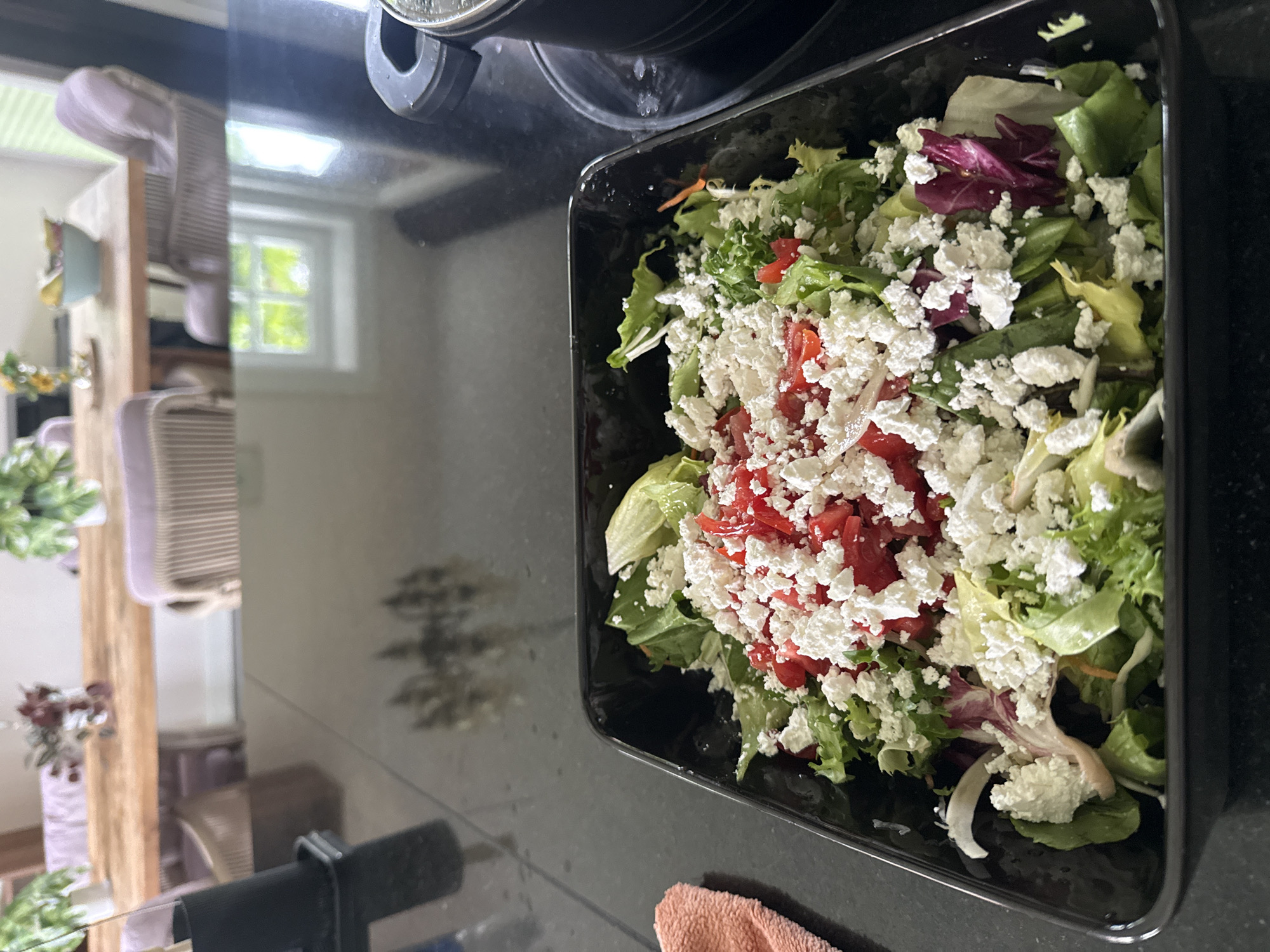 The image depicts a prepared salad in a black, rectangular serving dish, positioned on a dark, highly reflective countertop within a residential interior in Eyendorf, Germany.
The salad consists of a mixed green base, including light green and darker red/purple lettuce varieties, topped with a substantial amount of diced red tomatoes and generously sprinkled crumbled white cheese, consistent with feta. A slight sheen on the lettuce suggests the presence of dressing.
To the immediate left of the serving dish, a portion of a pinkish-orange fabric, possibly a kitchen towel, is visible. On the far right, a black cooking pot with a dark handle rests on a black surface, likely an induction cooktop.
The reflective countertop prominently displays inverted and somewhat distorted reflections of the background, which reveals a dining area. This area features a large, light-toned wooden dining table surrounded by multiple chairs with woven backs and light purple fabric cushions. Decorations on the table include two small potted green plants in white containers positioned at opposite ends, a clear vase with yellow flowers, a light blue bowl containing light-colored fruit, and another clear vase holding dark red decorative elements. In the background, a multi-pane window allows natural light into the room, revealing green foliage outside. The walls appear light-colored.
No people are visible in the frame, and no specific actions or interactions beyond the static arrangement of objects are depicted. The scene suggests a prepared meal in a domestic setting.
