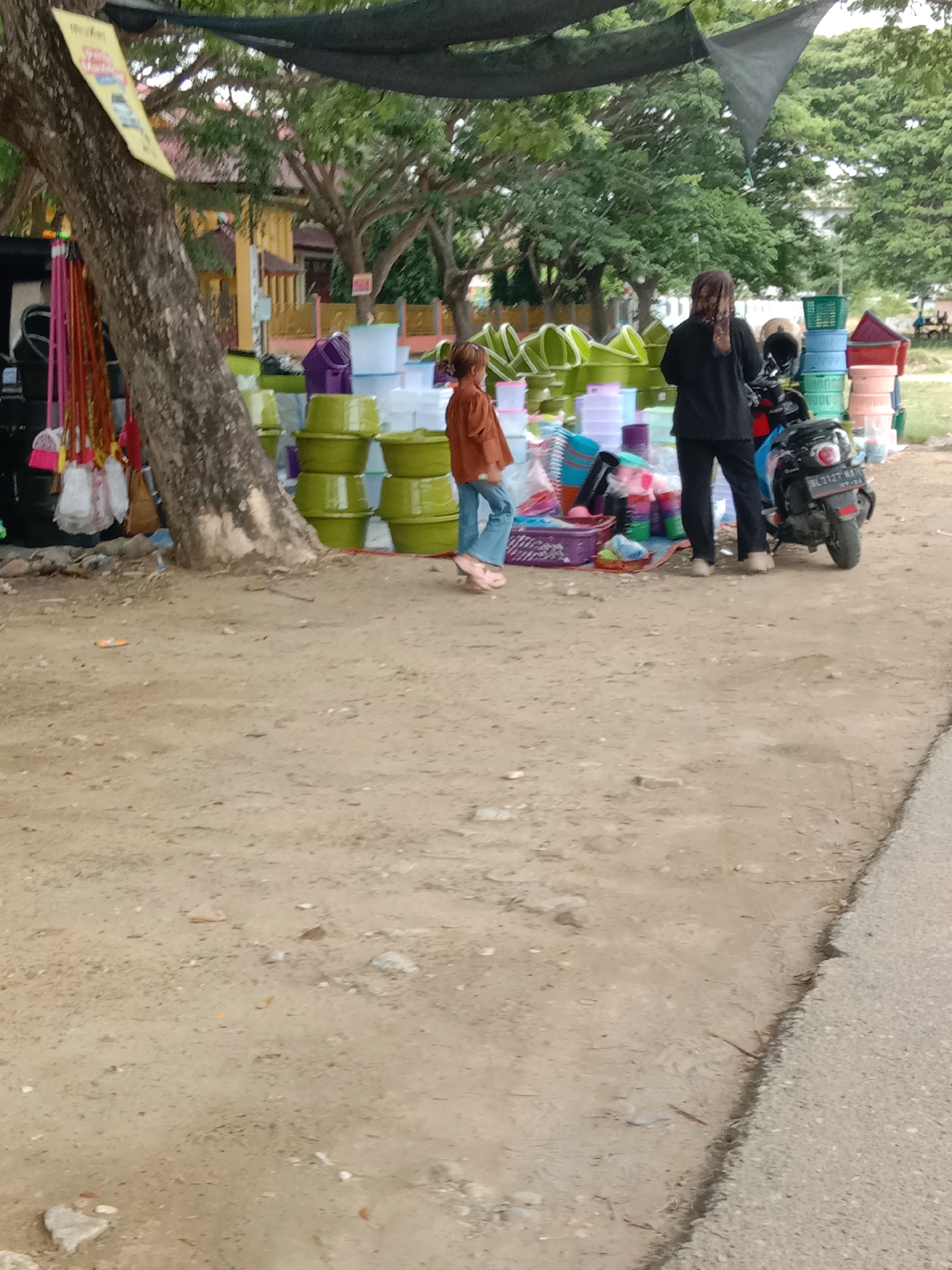 This image depicts an outdoor market scene, likely a roadside stall, in Lhokseumawe, Indonesia. The foreground is a dusty, unpaved ground surface next to a paved road on the right. In the midground, a variety of household items, primarily plastic containers and buckets in shades of green, purple, and white, are stacked and displayed. Some brooms and brushes are also visible, hanging from a tree trunk on the left.
Two individuals are present. A young person wearing a brown long-sleeved shirt and blue jeans stands facing left, looking at the merchandise. Behind them, a person wearing black clothing and a headscarf stands by a parked motorcycle, also facing away from the camera. The motorcycle is dark-colored with visible license plate details.
The background features trees with dense green foliage and buildings with yellow and red accents, suggesting a semi-urban or suburban environment. A black mesh-like covering is stretched above the market stall, providing shade. The overall impression is of a casual, everyday marketplace.