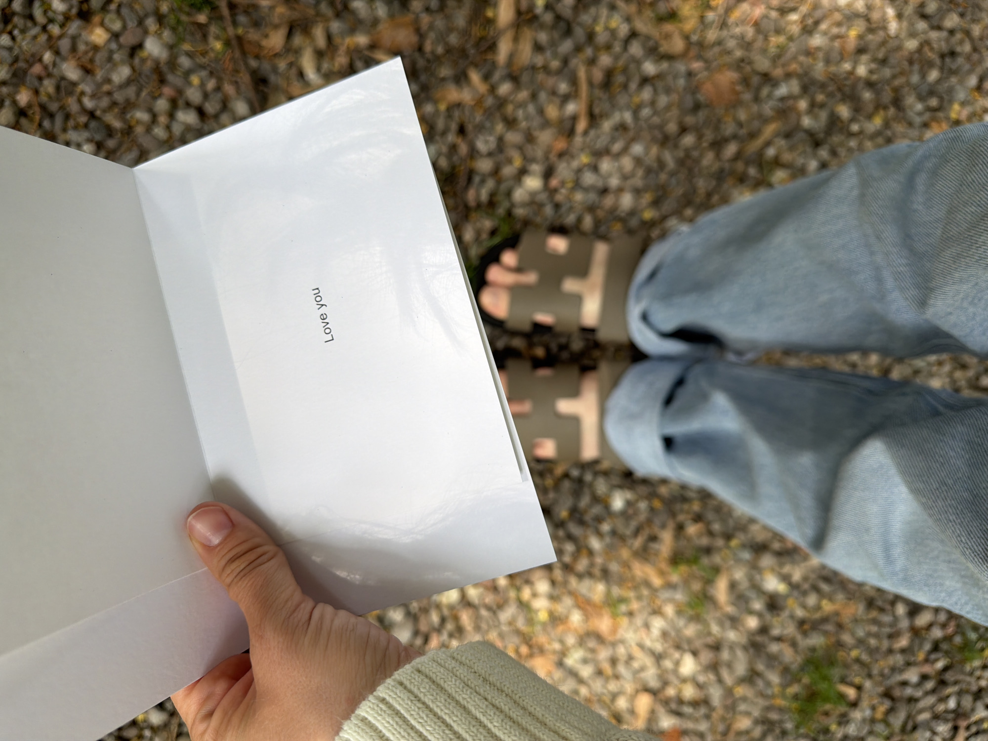 A close-up, overhead shot captures a person holding open a white greeting card with the words "Love you" printed inside. The person's hand, wearing a cream-colored, ribbed sweater, is visible on the left side of the frame, holding the card. The lower half of the image displays the person's legs clad in light-wash denim jeans, cuffed at the bottom, and olive-green slide sandals. The ground is covered in small, light-colored pebbles and scattered autumn leaves, suggesting an outdoor setting. The lighting is natural and diffused, casting soft shadows. The scene is likely located in a park or a garden area, possibly in the city of Penzano, Italy, given the stony terrain and foliage.