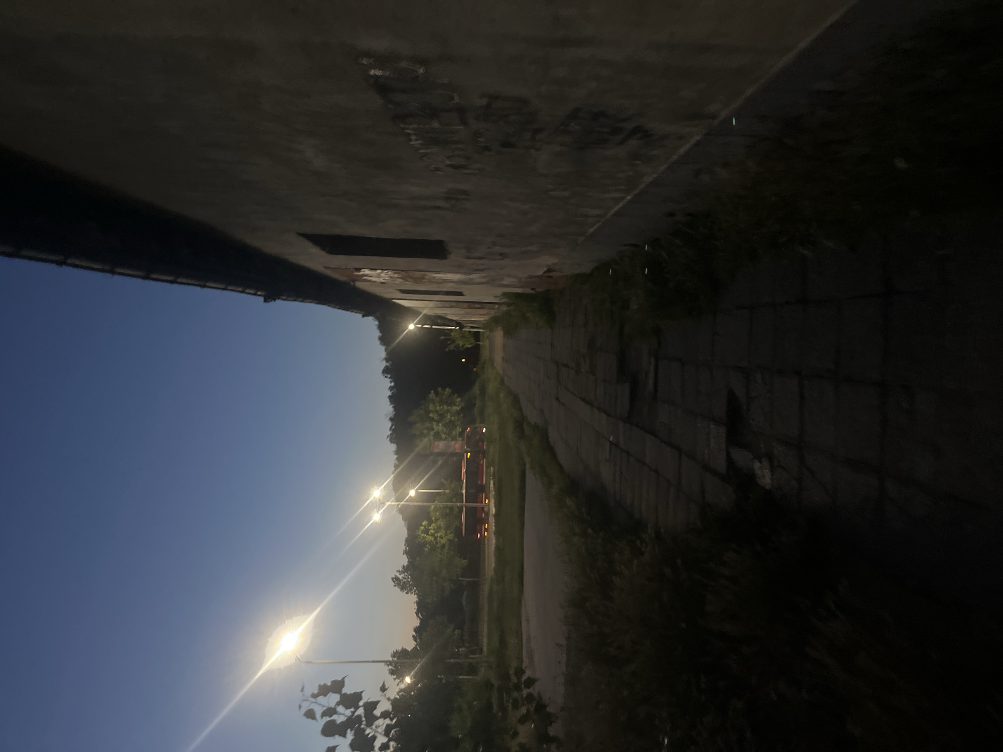 The image shows a street scene at dusk, likely in Plovdiv, Bulgaria. A narrow, paved walkway runs along the right side of the frame, adjacent to a long, weathered building with graffiti on its exterior. The building's wall is textured and appears to be made of stucco. Across the walkway, on the left side of the image, is an open area with patchy grass and several tall lampposts that illuminate the scene. In the distance, a red bus is visible, parked or driving in the middle ground, surrounded by trees and foliage. The sky is a deep blue, characteristic of twilight. The lighting from the lampposts creates a dramatic contrast, casting long shadows and highlighting the texture of the walkway and the building. The overall impression is one of an urban fringe area, possibly an industrial zone or the outskirts of a neighborhood, captured during the transition from day to night. There are no people visible in the image.