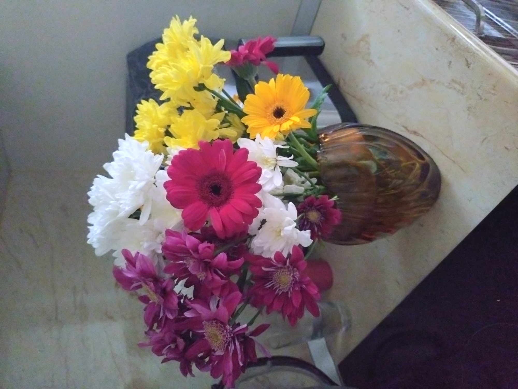 A vase of colorful flowers sits on a marble-like countertop. The vase, made of dark glass, lies on its side. The flowers include yellow, white, pink, and burgundy-colored varieties, some of which appear to be chrysanthemums and gerbera daisies. The countertop has visible patterns. The setting is a domestic interior. There is no indication of the location being Stara Zagora, Bulgaria.