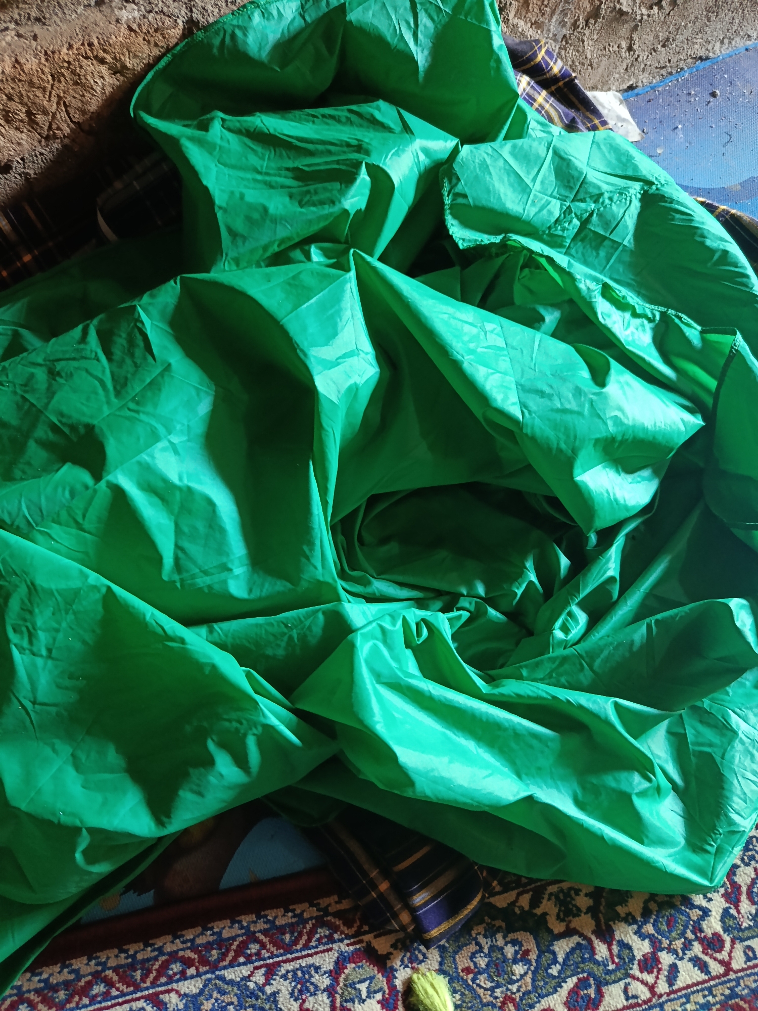 A crumpled, bright green tarp is spread out on a patterned rug and a blue mat, next to a rough, brown wall. The tarp is creased and folded, with sections of light and shadow across its surface. In the upper left, a textured, earthy brown wall is visible. Below it, a blue mat with a faint speckle pattern can be seen. The lower portion of the image is dominated by a rug with a detailed geometric pattern in red, blue, and cream. A small piece of yellow yarn is visible on the rug in the bottom center. The scene appears to be an indoor, possibly storage or makeshift area. The location in Karangmojo, Indonesia is not visually evident from the image.