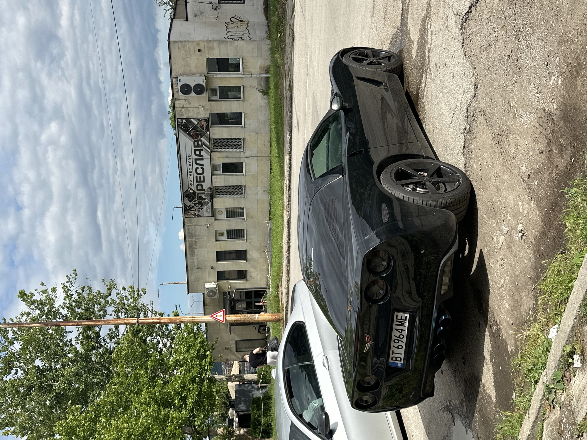 A black Chevrolet Corvette is parked on a cracked asphalt surface outside a building. The car's rear is facing the camera, and its license plate reads "BT 6964 ME". A silver car is parked to the left of the Corvette. The building behind the cars has a sign that reads "ФИТНЕС КЛУБ ПРЕСЛАВ". A man in a black t-shirt and white shorts is walking away from the building, carrying a black bag. A utility pole with a red and white triangular sign stands between the cars and the building. The scene is under a cloudy sky.