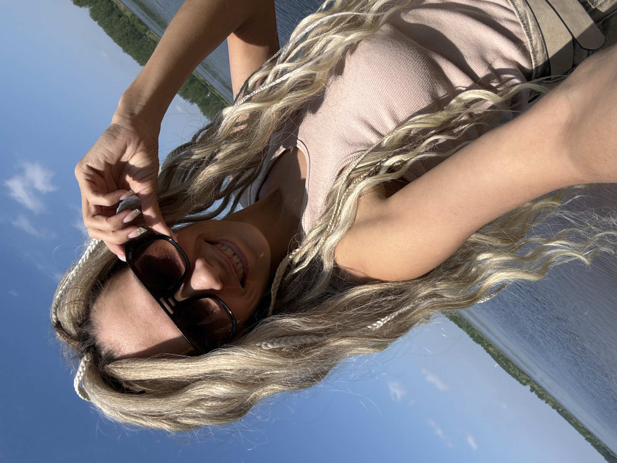 A low-angle outdoor shot features a woman with long, blonde, braided hair wearing sunglasses and a light brown top. She is smiling and holding the sunglasses with her left hand. The background shows a body of water and a green shoreline under a blue sky. The location appears to be in a sunny, outdoor setting.
