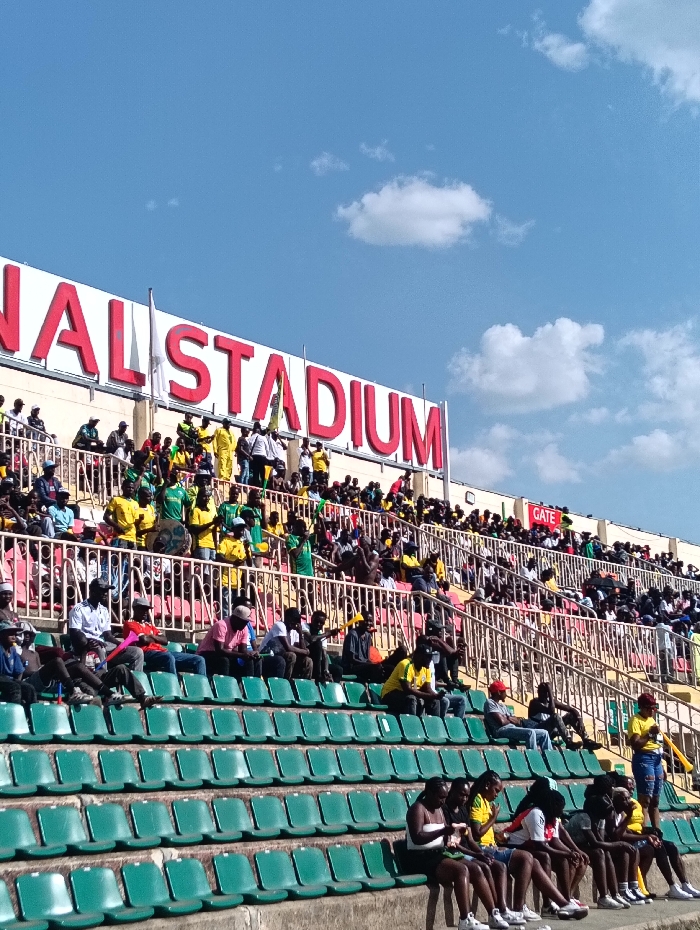 The image displays a section of a stadium grandstand under a bright blue sky with scattered white cumulus clouds. A prominent sign with large red lettering on a white background reads "NATIONAL STADIUM", indicating the location is a national stadium in Nairobi, Kenya. The grandstand structure consists of concrete tiers fitted with rows of green plastic bucket seats; the lowest rows in the foreground are largely empty.
Multiple individuals, predominantly young adults, are present. The upper tiers are densely populated, with many spectators wearing yellow and green attire. Some in this section are standing, while others are seated. One individual in the upper-mid section is depicted holding what appears to be a drum. The middle tiers show fewer, more sparsely distributed individuals seated in casual positions, wearing a mix of yellow, red, and dark clothing. In the immediate foreground, a row of approximately eight individuals, mainly young females, are seated on the green chairs, dressed casually in shorts, t-shirts, and sneakers; some are looking downwards.
A smaller red sign with white text "GATE" is visible on an upper wall to the right of the main "NATIONAL STADIUM" sign. A white flag is affixed to a flagpole positioned near the "NATIONAL" segment of the stadium sign. The scene is illuminated by strong daylight, casting visible shadows.