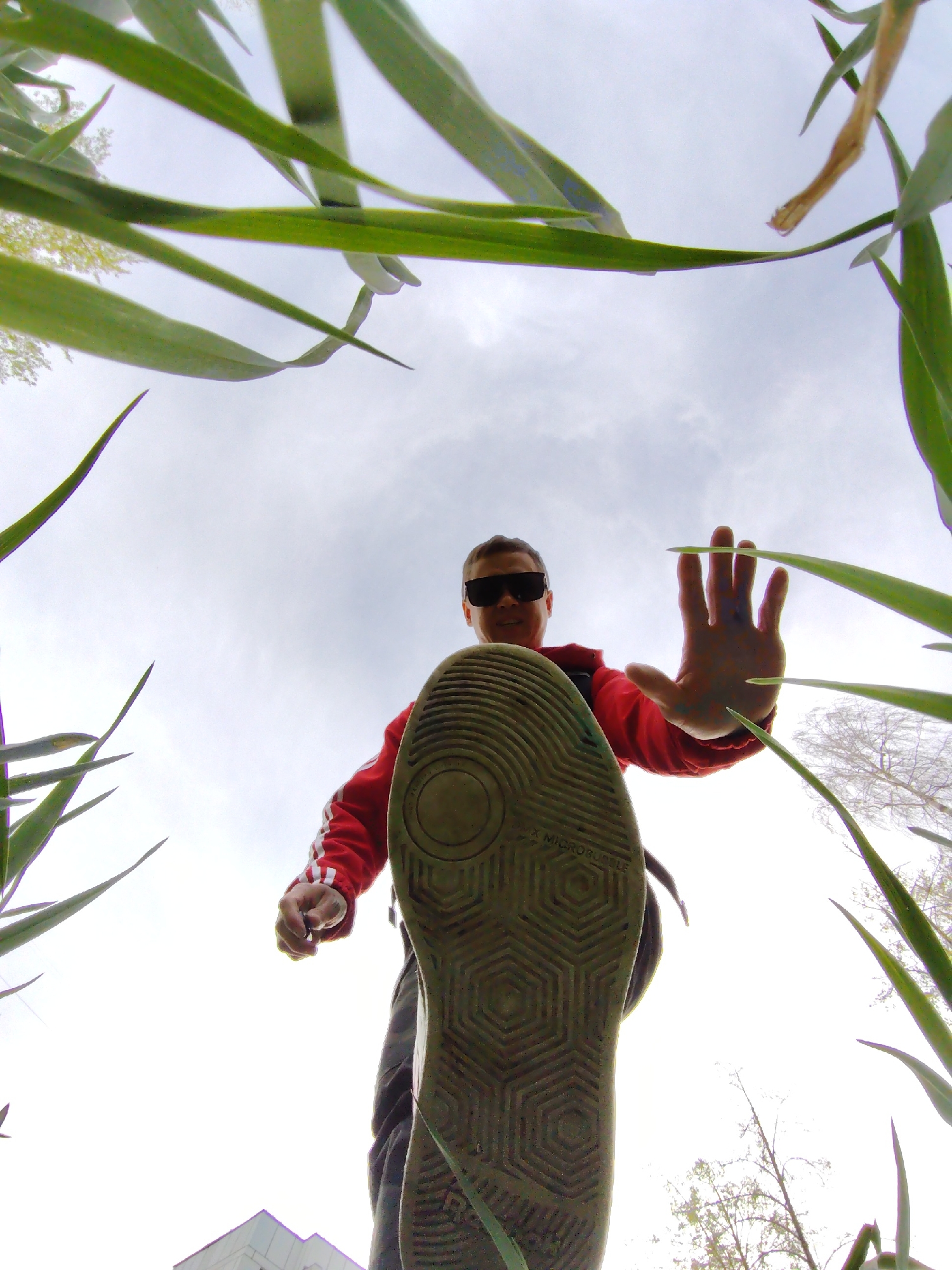 The image depicts a low-angle perspective looking up at a person standing over the camera. The person is wearing sunglasses, a red tracksuit jacket with white stripes, and gray pants. The sole of their shoe is prominently displayed, obscuring the lower part of their body. Their right hand is raised in a "stop" gesture. The background consists of green grass blades reaching up and an overcast sky. In the distance, a building can be seen on the left, and trees with sparse foliage on the right. The text "X MICROBUBBLE" is visible on the shoe sole. The location is in the city of Chaikovsky, Russia.