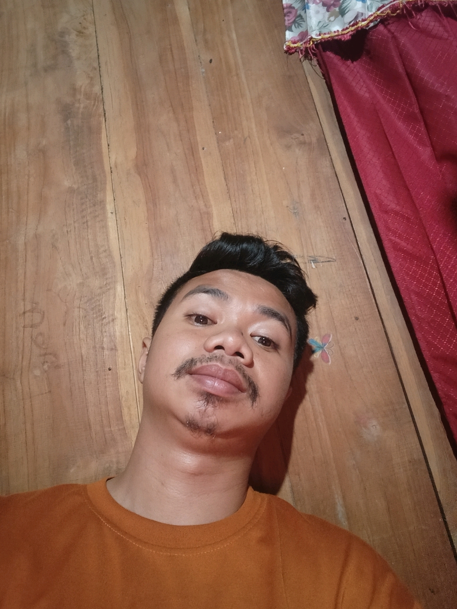 A young male with dark hair, mustache, and goatee is shown from the chest up, looking at the camera from a slightly upward angle. He is wearing an orange t-shirt. The background is a wooden surface, possibly a floor or wall, with visible wood grain and knots. To the right, a red curtain with a floral pattern at the top is visible. A small, colorful butterfly sticker is affixed to the wooden surface near the man's head. The image appears to have been taken indoors in a residential setting in Galuk, Indonesia.