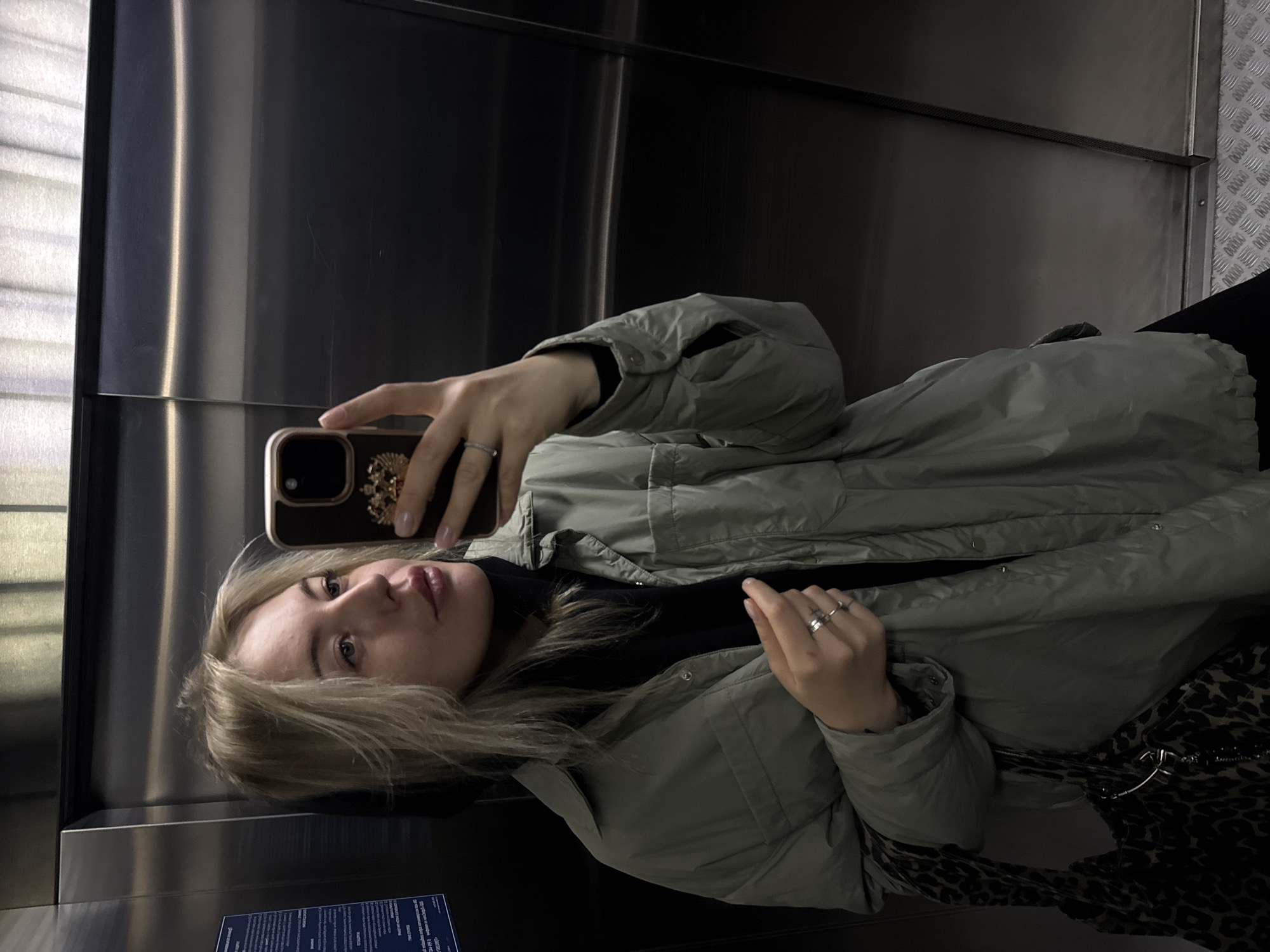 A woman with blonde hair is taking a selfie in an elevator. She is wearing a light green puffer jacket over a black top, and a leopard print bag is visible. The elevator has stainless steel walls. A sign with Russian text is visible in the lower left corner of the elevator. The location is Krasnoyarsk, Russia.