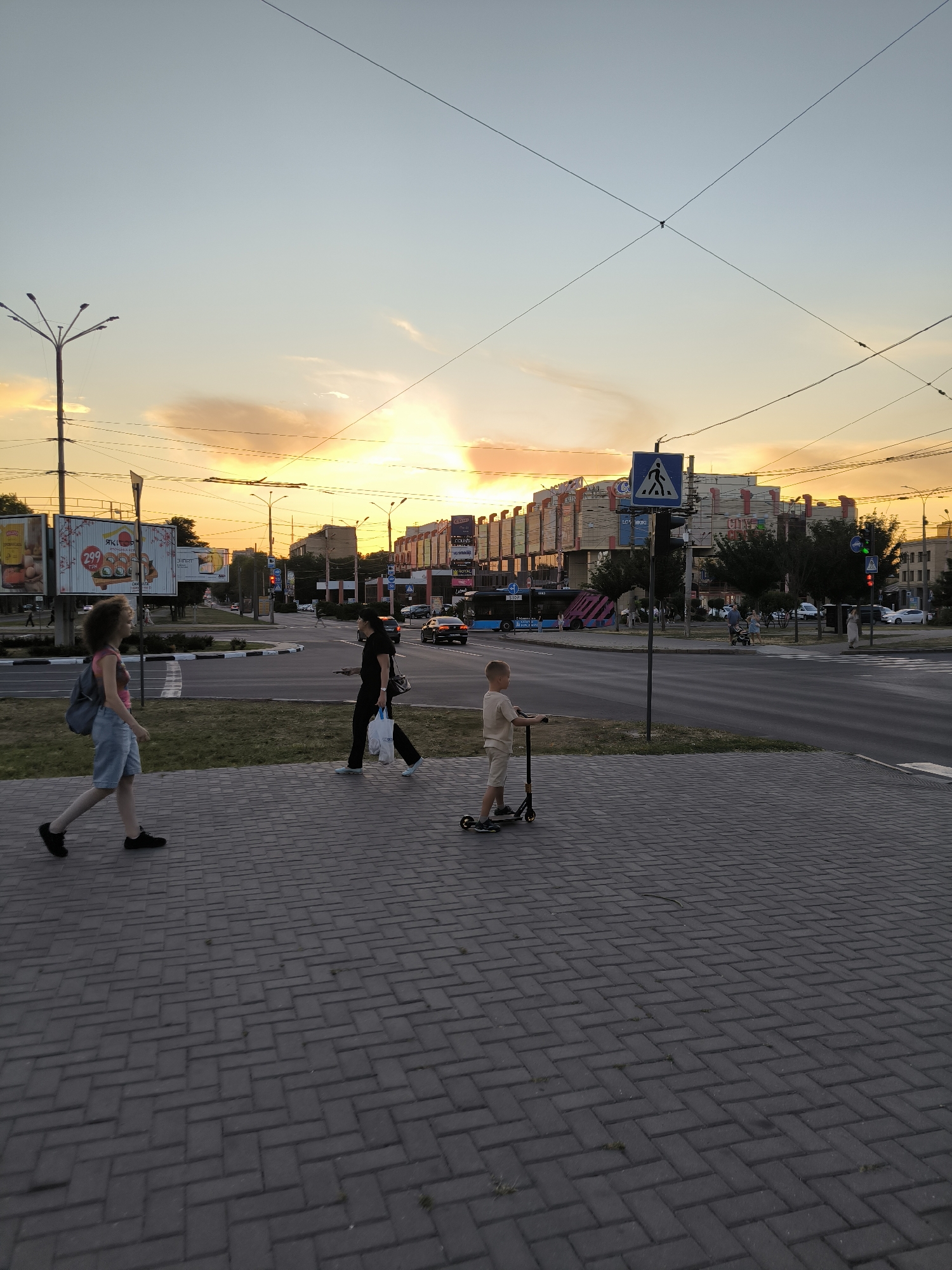 The image shows a sunset scene on a street in Mykolaiv, Ukraine. In the foreground, a young boy on a scooter rides along a paved sidewalk. Further down the sidewalk, a woman in dark clothing walks with shopping bags. To the left, a young woman with curly hair walks with a backpack. Beyond the sidewalk, a street with traffic is visible, including cars and a bus. Buildings line the street, and a pedestrian crossing is marked. Overhead, power lines crisscross the sky. The setting sun casts a warm glow over the scene, illuminating the clouds in the sky.