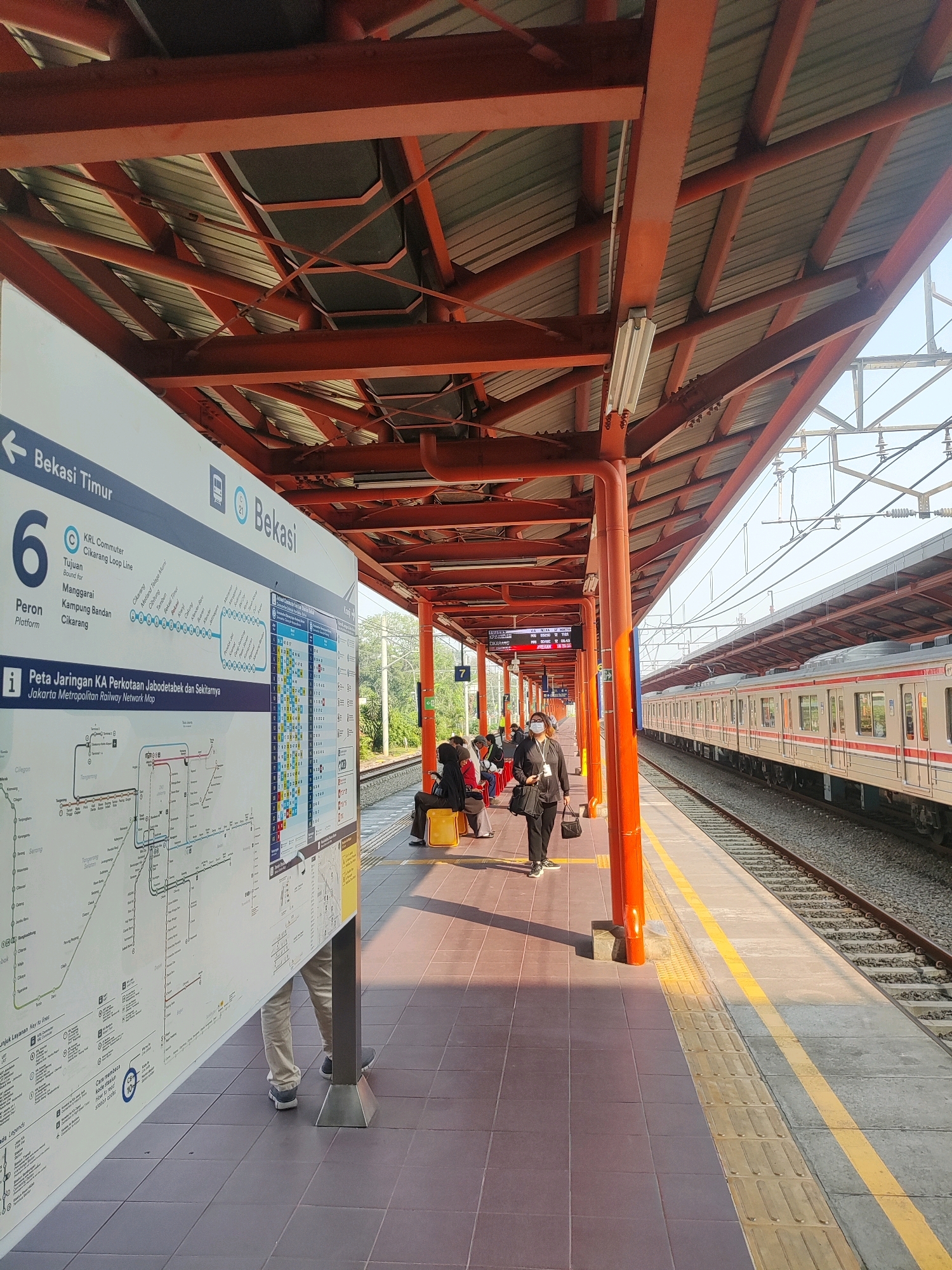 The image depicts a train platform in Bekasi, Indonesia, specifically at a station serving the KRL Commuter Line. A train with a red and cream livery is stopped at the platform on the right side of the frame. On the left, a large informational display board shows a map of the Jakarta Metropolitan Railway Network. Several people are visible on the platform. Two individuals are standing near the informational board, and a group of people are seated on yellow benches further down the platform. The platform is covered by an orange, metal-framed roof supported by red pillars. The ground is tiled with dark grey and brown tiles, with a yellow tactile warning strip along the platform edge. The sky is clear and bright, suggesting it is daytime. The overall scene is typical of a commuter train station environment.