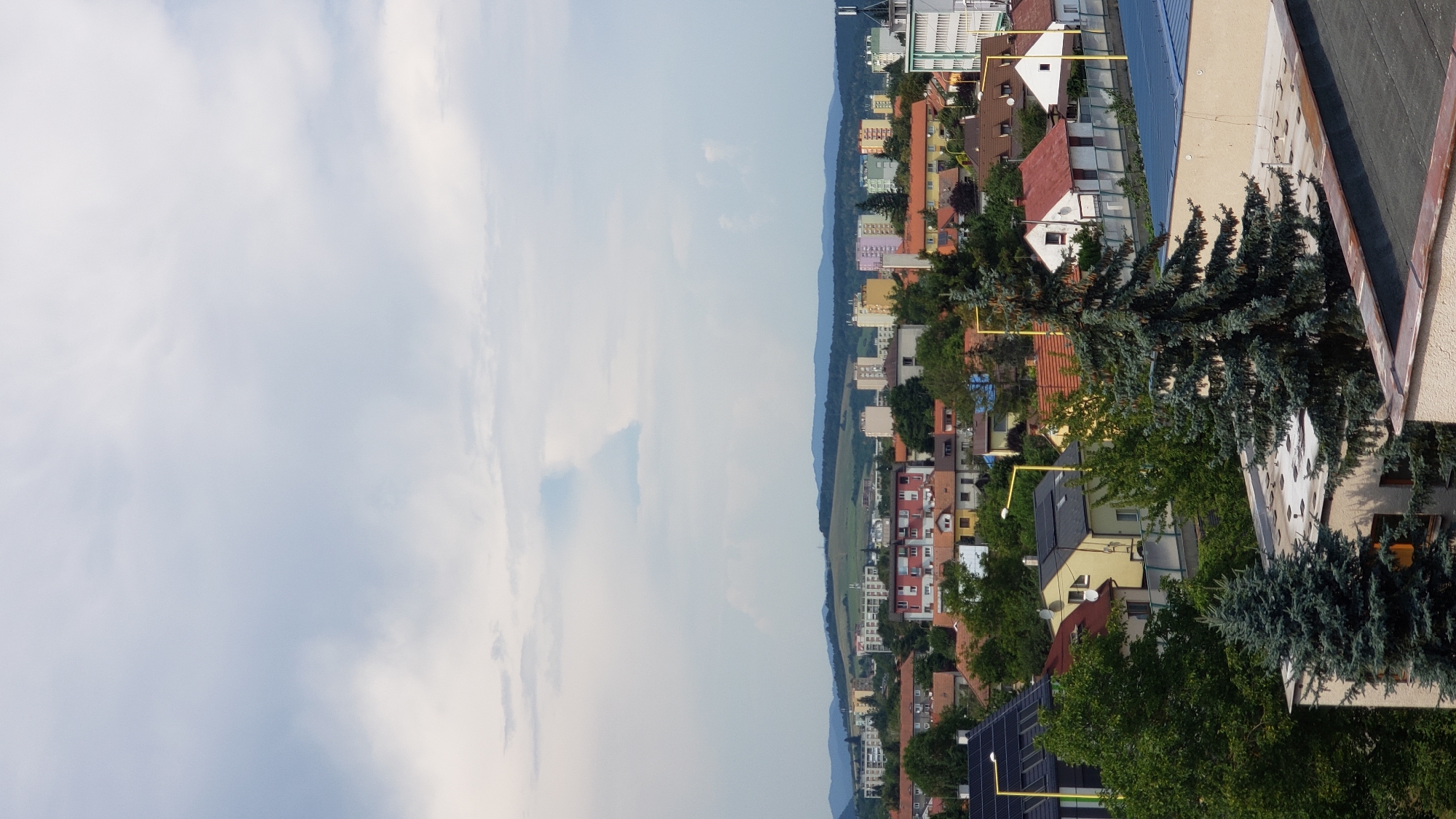 The image depicts an aerial view of a cityscape in Prešov, Slovakia, under a cloudy sky. In the foreground, rooftops of residential buildings are visible, some with solar panels. Large evergreen trees stand prominently, their branches reaching towards the sky. Beyond the foreground, a denser collection of houses with red-tiled roofs is seen, interspersed with trees. Further in the distance, apartment buildings of varying heights and colors, including beige, light yellow, pink, and light green, form a distinct section of the city. A prominent hill with green fields and a dark structure on its crest rises behind the apartment buildings, and rolling hills form the backdrop under the overcast sky. The scene captures a residential area of Prešov, likely a neighborhood with a mix of single-family homes and multi-story apartment blocks, set against a natural landscape.