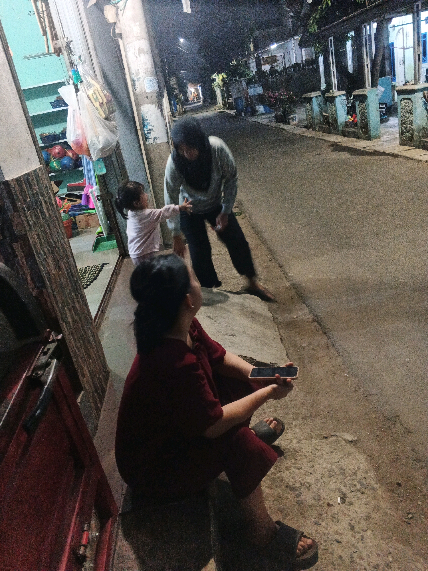 In Kadu, Indonesia, at night, a woman in a maroon top and dark pants sits on a concrete step holding a smartphone. To her left, a young child in a pink shirt reaches out towards a woman in a grey top and hijab. The woman in the grey top is looking down at the child. Behind them, a street is visible with buildings on either side. To the left of the seated woman, there appears to be a shop entrance with shelves containing various items. The overall environment suggests a residential or commercial street scene at dusk or night.
