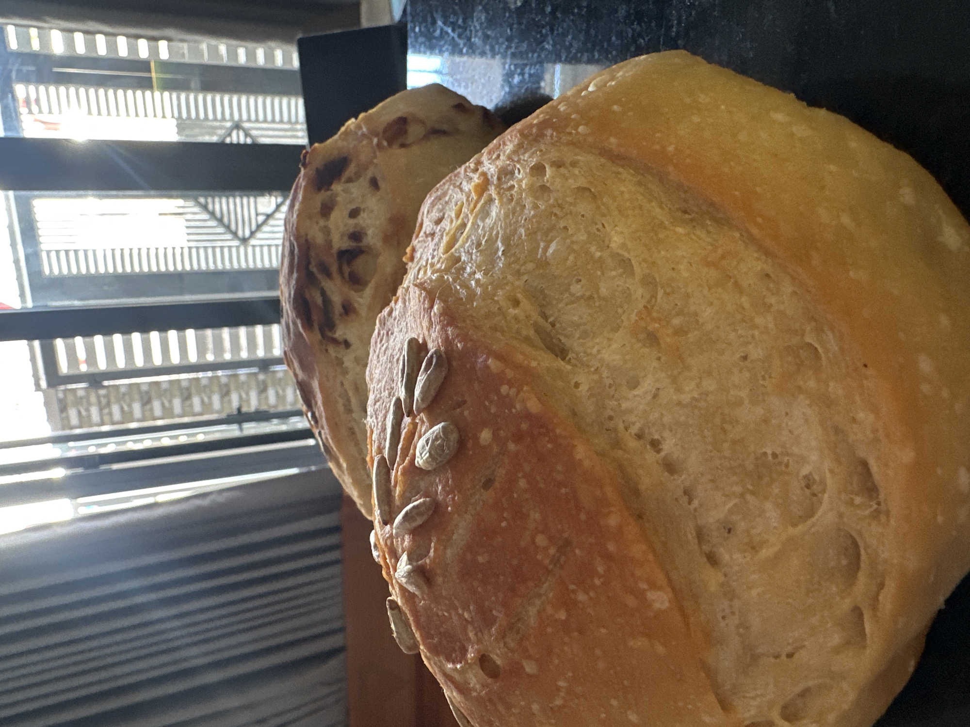 The image shows two loaves of bread. The loaf in the foreground has a light golden crust with sunflower seeds embedded on its surface. The second loaf is behind the first and has a darker, more porous surface. The background includes a window with horizontal slats and a dark countertop surface. The location is potentially inferred based on the city of Bac Nha Trang Ward, Vietnam, but there are no visual cues from the image itself that definitively confirm this location. There is no presence of human activity.