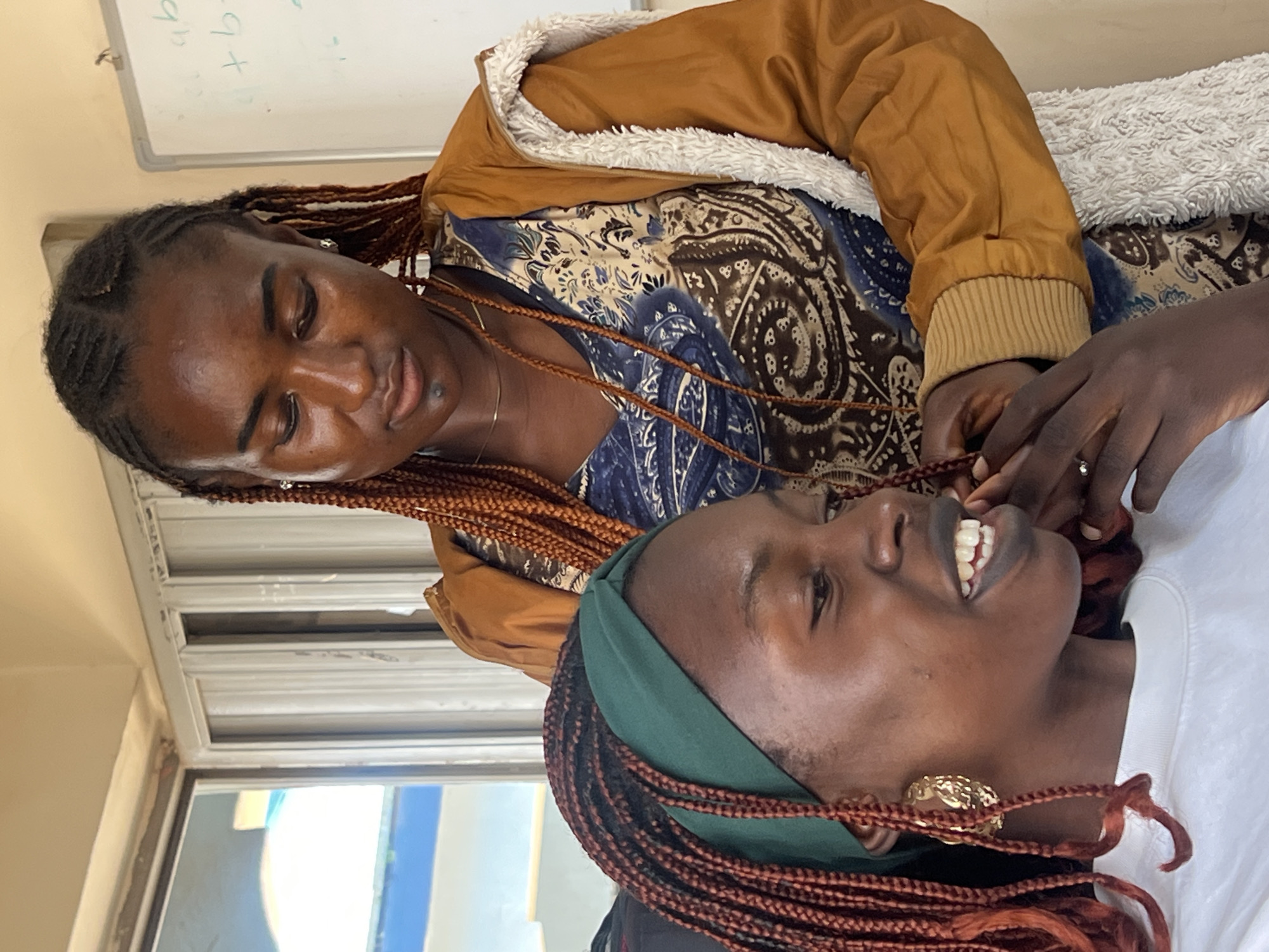 The image shows two young women with braided hair indoors, likely in a classroom or office setting in Jos, Nigeria. The woman in the foreground, wearing a green headband and a white sweatshirt, is smiling and looking to her left. She has large, ornate gold earrings. The woman behind her, wearing a patterned shirt and a brown jacket with a furry lining, is looking down at the woman in front and appears to be adjusting her hair. In the background, there is a window with a view of a blue structure and a whiteboard with some writing on it. The lighting suggests it is daytime.