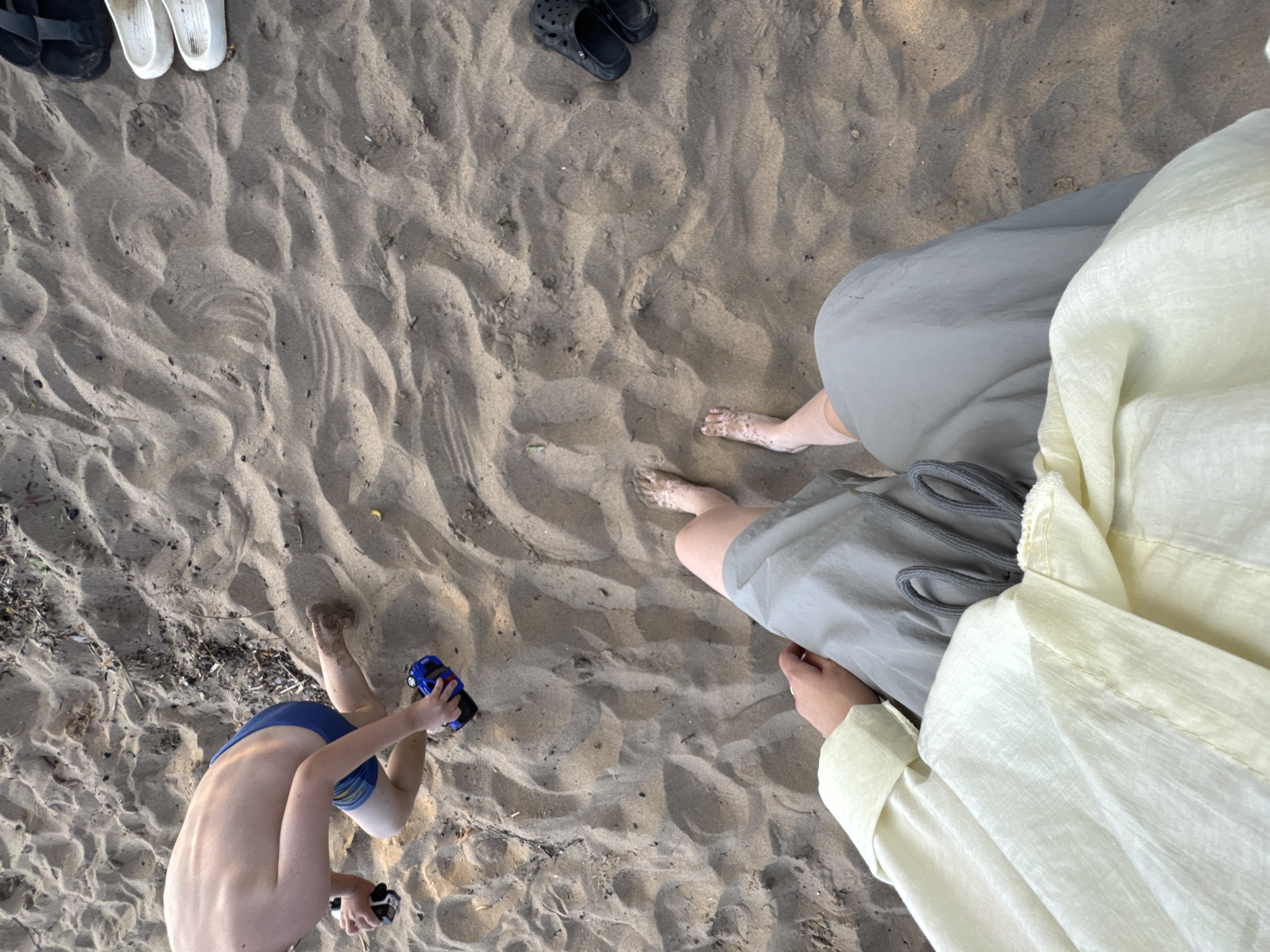 The image shows a high-angle view of a sandy beach with several people. Two individuals are partially visible, wearing light-colored clothing, possibly a shirt and shorts, and their legs are seen resting on the sand. Another person, a child wearing blue swim trunks, is seated on the sand, holding a small blue toy car and a black object. They appear to be playing on the beach. In the background, several pairs of sandals and shoes are placed on the sand, including white slides and black clogs. The sand has distinct wavy patterns, suggesting it has been shaped by wind or water. The lighting suggests it might be late afternoon or early morning, casting soft shadows. The overall scene depicts a relaxed day at the beach. The location is Chaikovsky, Russia. No visible text is present in the image.