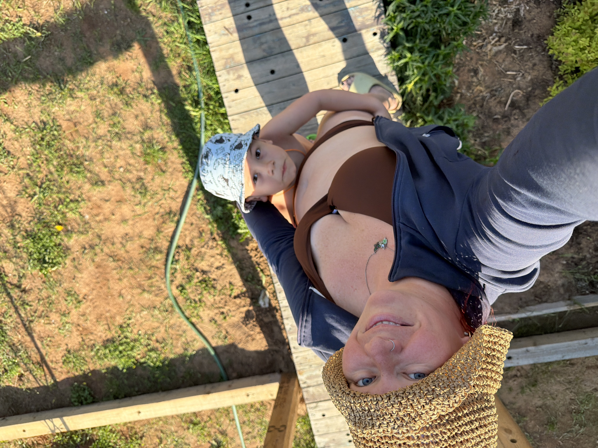 A selfie taken from above shows a woman wearing a brown bikini, a dark blue jacket, and a tan woven hat holding a young child wearing a blue bucket hat with white patterns. They are standing on a wooden walkway with gaps between the planks, which is surrounded by green vegetation and bare earth. A green garden hose is also visible in the image. The location is not discernible as Chaikovsky, Russia based on the content of the image.