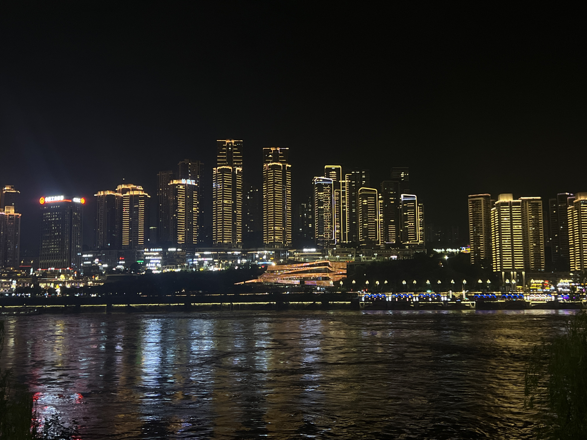 The image captures a nighttime cityscape of Jiangbeicheng Street in China, dominated by a waterfront illuminated by numerous buildings and their reflections in the water. A wide body of dark water, likely a river, occupies the foreground, with the lights from the city reflecting on its surface, creating shimmering patterns of yellow, white, blue, and red. Along the waterfront, a line of buildings and structures are brightly lit.
In the background, a dense collection of skyscrapers forms the city skyline. These buildings are illuminated with a warm yellow light, highlighting their architectural details and vertical lines. Some buildings feature distinct patterns of light, such as horizontal bands or vertical stripes. One prominent building in the mid-ground appears to have a unique, layered, and angular design, also lit with warm colors.
Further along the waterfront, a row of boats or barges is visible, each adorned with decorative blue and white lights. A long, thin band of lights runs along the edge of the landmass, separating the water from the buildings. The overall impression is one of a vibrant and bustling urban environment at night, characterized by extensive artificial lighting. The sky above is a dark, featureless expanse. There are no discernible people or specific actions taking place. The focus is on the architectural lighting and the reflection on the water.