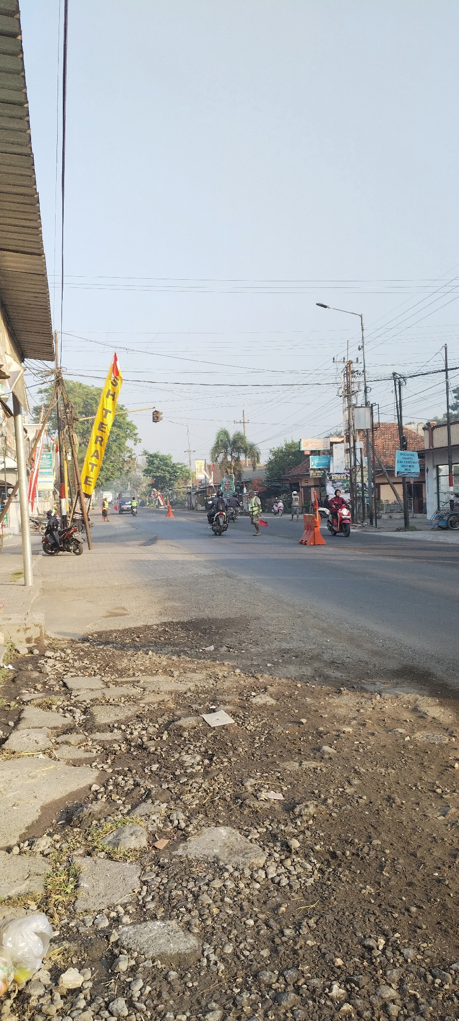 The image depicts a street scene in Jombang, Indonesia. A yellow banner with red lettering that appears to read "SH TERATE" hangs vertically from a utility pole. In the foreground, the road is uneven and appears to be under repair or in disrepair, with exposed gravel and broken pavement. Several motorcycles are present, some parked and others in motion on the asphalt road. A few individuals are visible, some wearing reflective vests and holding red flags, possibly directing traffic or indicating a road closure. Orange traffic cones are placed on the road. Buildings line the street, some appearing to be shops or residences. Utility poles with numerous wires crisscross the sky, which is clear and pale. The overall impression is a busy, functional street, possibly experiencing temporary disruption due to road work or an event.