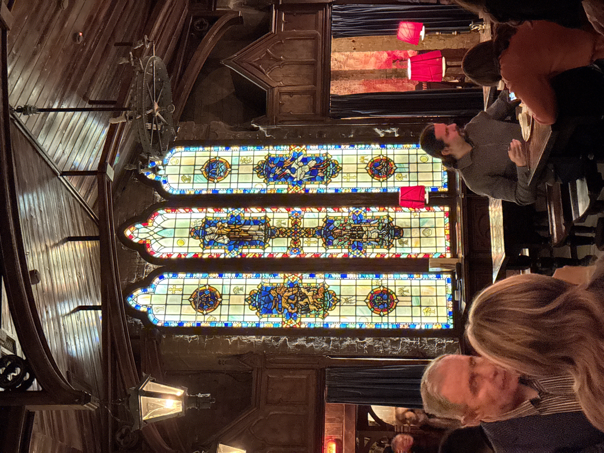 The image depicts an interior scene with a large, ornate stained glass window as a prominent feature. The window is divided into three horizontal sections, each filled with intricate designs and vibrant colors, predominantly blues, yellows, and reds. The surrounding architecture suggests a building with a historical or religious significance, with dark wood paneling and decorative molding visible.
Several people are present in the scene, appearing to be patrons of what might be a restaurant or bar. They are seated at tables, engaged in conversation or looking at their phones. A few red pendant lamps are hanging nearby. The atmosphere appears dimly lit and somewhat intimate.
While the image itself does not provide explicit information about the location being in Moscow, Russia, the architectural style, the design of the stained glass window, and the overall ambiance could be indicative of a historical building repurposed into a commercial space, a trend seen in many cities including Moscow. However, this is only suggestive, not definitive, without additional context.