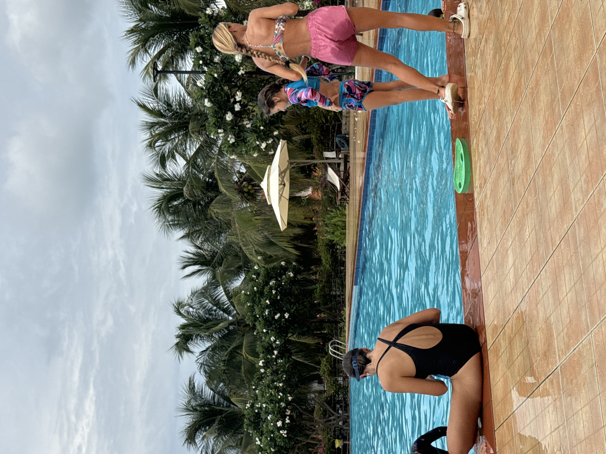 The image depicts an outdoor swimming pool scene under an overcast sky, surrounded by lush tropical vegetation. The perspective is tilted, showing the pool running horizontally across the frame.
In the foreground, a person wearing a black one-piece swimsuit and swimming goggles is bent over at the edge of the blue swimming pool, possibly about to enter the water or reach for something. To their right, the pool deck is covered with light brown and orange tiles. A green swimming kickboard floats in the water near the edge.
Further back, two other individuals are standing by the pool's edge. One, an adult or older child with blonde hair styled in a braid, wears a pink bikini top and pink shorts. Beside them, a younger child with dark hair is visible, wearing a long-sleeved, colorful patterned swimsuit. White shoes are seen near their feet on the tiled deck.
The background is dominated by numerous palm trees and bushes with white flowers, indicative of a tropical environment. A light-colored umbrella is partially visible amidst the foliage. The sky is largely covered with light grey clouds, suggesting an overcast day. The scene evokes a recreational and relaxed atmosphere, likely at a resort or private residence in a warm climate, consistent with the provided context of Phu Thuy Ward, Vietnam. No discernible text is visible in the image.