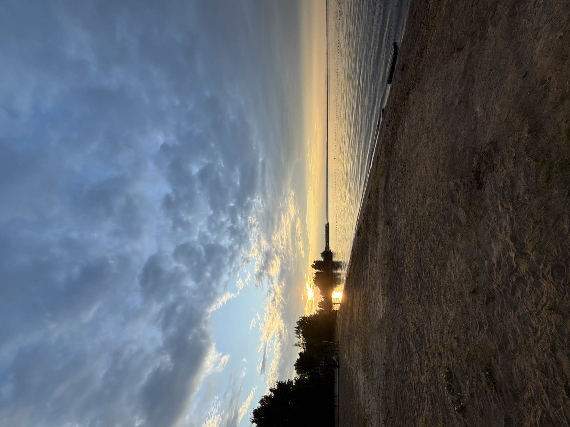 The image depicts a serene beach scene at sunset on the Kama River embankment in Chaikovsky, Russia. The sky is filled with dramatic clouds, with streaks of sunlight breaking through, casting a warm glow over the water and the sand. The sun, low on the horizon, is visible between the silhouetted trees on the left side of the frame. A few scattered people are present on the sandy beach, some walking near the water's edge, while others are further back, silhouetted against the setting sun. A small wooden structure or play area is visible among the trees on the left. The water is calm, with gentle ripples reflecting the sky. A marker buoy line extends across the river, and a dark, rectangular object, possibly a small raft or dock, is partially submerged near the shore on the right. The foreground consists of textured sand with some sparse vegetation.