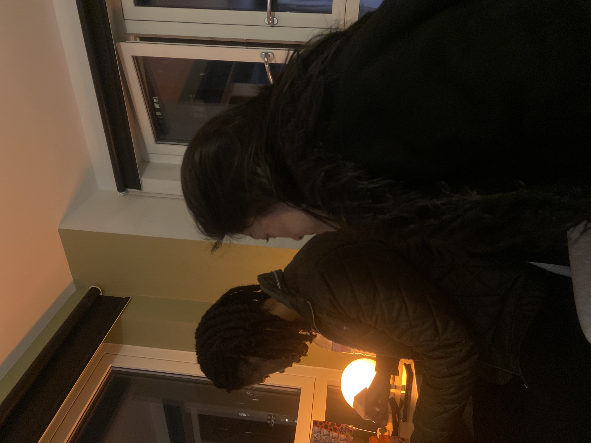 The image displays an indoor scene, captured in what appears to be a vertical orientation (if the subjects were standing upright), showing two individuals in a dimly lit room.
On the left side of the frame, a white-framed window is visible, partially obscured by a black roller blind that extends vertically. Another window is also discernible in the background. The wall to the left of the window is a light, neutral color, while the wall further right adopts a light yellowish-green hue.
Two people are present. In the foreground, a person with long, dark hair, wearing a dark coat or jacket, is seen in profile, looking downwards. Their upper body is partially visible. In front and slightly to their right, another individual with dark dreadlocks or braids is also seen in profile, facing towards the right and looking downwards. This person is wearing a dark, possibly quilted, jacket.
On the right side of the image, a spherical lamp emits a warm orange glow, illuminating its immediate surroundings. Below the lamp, some indistinct objects are placed on a surface, possibly a table or shelf.
The scene suggests a quiet, possibly intimate, moment between the two individuals. The dark exterior visible through the windows, contrasted with the prominent warm artificial light from the lamp, indicates that it is evening or nighttime. No specific emotions are clearly discernible from their partially obscured faces, and no text is visible within the image. The provided location context is London, United Kingdom.