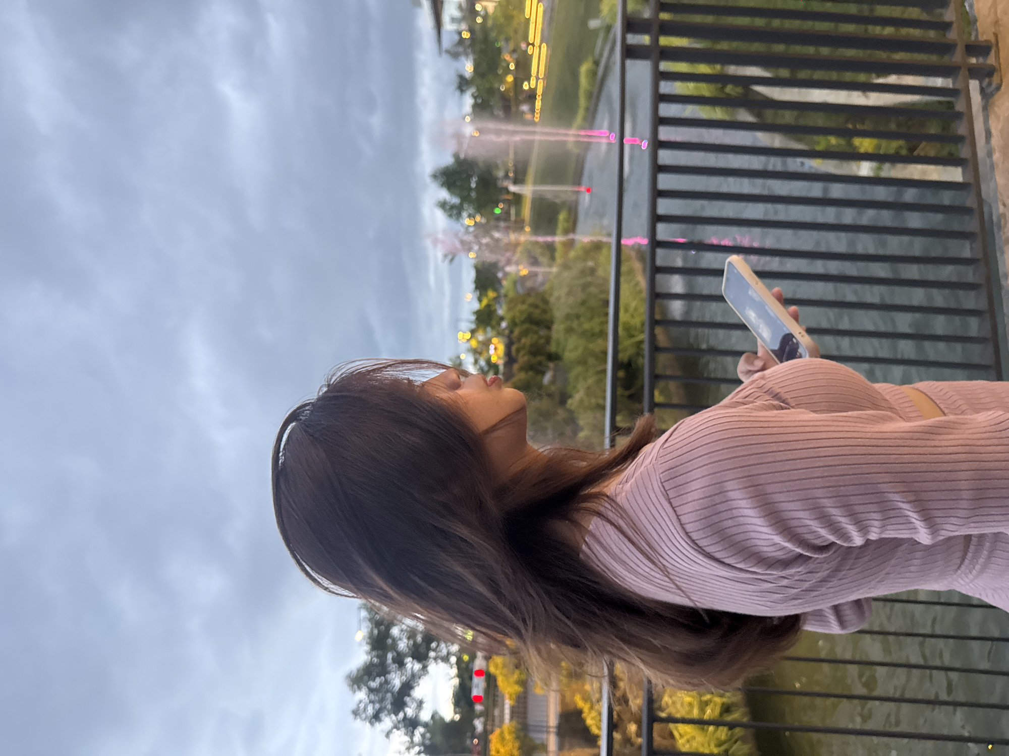 A young woman with long, dark brown hair stands outdoors in Davao City, Philippines, holding a cell phone, possibly taking a photo. She is wearing a light pink long-sleeved, ribbed shirt. She is positioned on a balcony or elevated platform with a metal railing. Behind the railing is a body of water with illuminated fountains. The sky is overcast. Trees and other greenery are visible in the background, along with indistinct city lights.