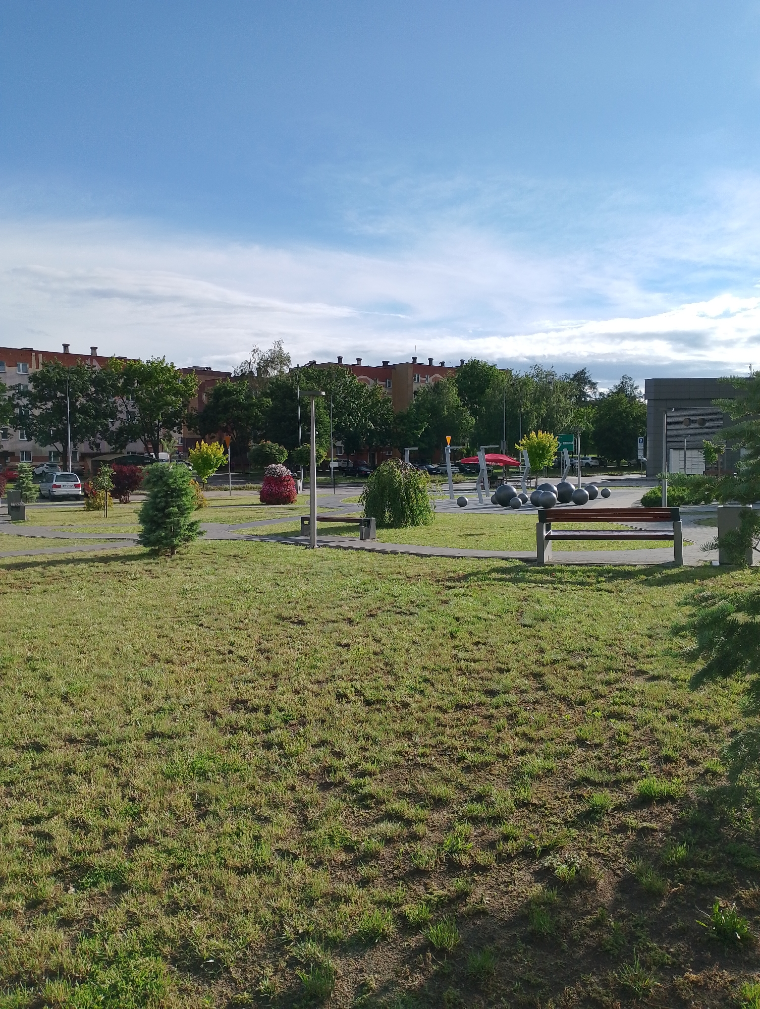 This is a daytime, eye-level, full-shot photograph of a park area in Kolno, Poland. The foreground is dominated by a grassy lawn with patches of dry, matted grass and sparse green growth. In the mid-ground, a paved pathway winds through the park. To the right of the pathway sits a wooden bench with a dark brown seat and grey concrete supports. Further into the mid-ground, a playground structure with red metal supports and grey spherical objects is visible. Several large, dark grey, spherical objects are arranged on the ground near the playground. To the left of the pathway, a small evergreen tree stands, and beyond it, a variety of bushes and trees are planted, including a vibrant red and pink floral display. In the background, a multi-story brick apartment building with a red-tiled roof and visible chimneys is situated behind a line of trees. Another modern building with dark cladding and a pitched roof is visible to the far right. The sky is a clear blue with scattered wispy white clouds. A silver car is parked on the road to the left of the park.