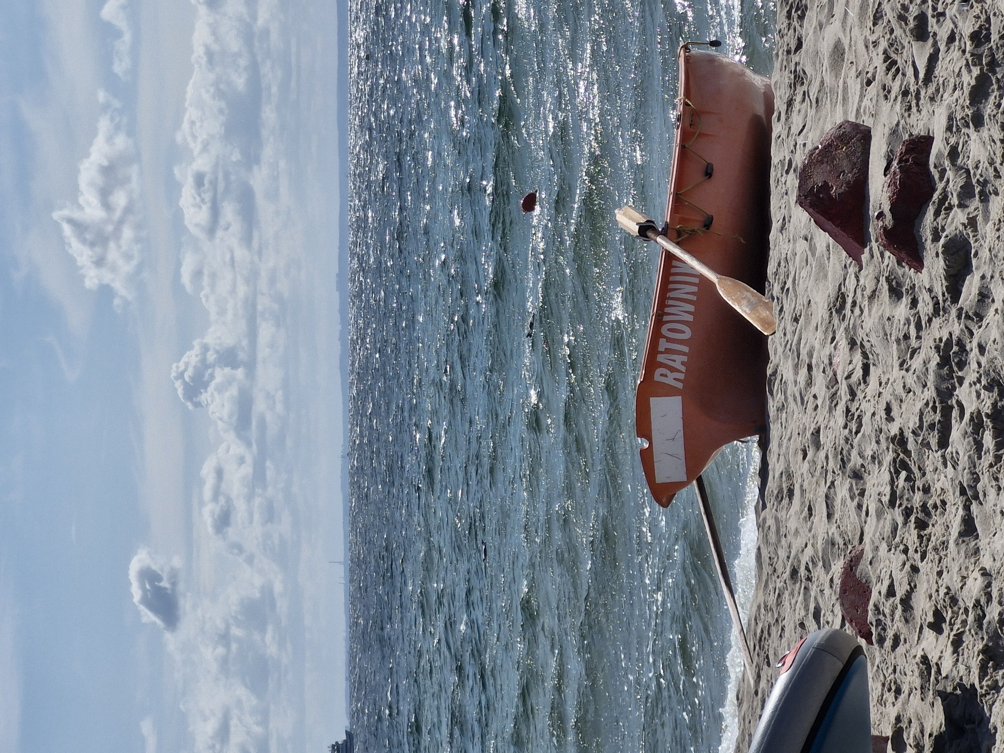 An orange lifeguard boat with the word "RATOWNIK" (meaning rescuer in Polish) written on its side rests on a sandy beach. A wooden oar is placed inside the boat, resting against its edge. The boat is positioned facing the slightly choppy, sparkling blue water, with a calm horizon line in the distance. The sky above is a pale blue with scattered white clouds. The beach itself is sandy with some small, dark red rocks and the edge of another grey inflatable object visible in the lower left corner. The scene captures a peaceful moment at the seaside, likely in Hel, Poland, given the context.